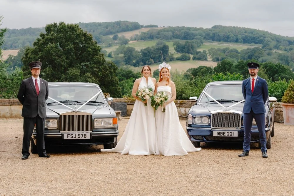 Two brides in wedding dresses holding bouquets, standing between two decorated vintage cars adorned with white ribbons, with two grooms in suits standing beside the cars, outdoors with green hills and trees in the background.