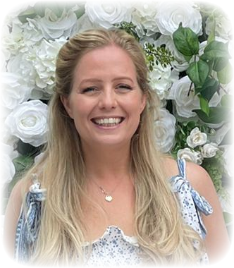 A woman with long blonde hair smiling in front of white flowers and green foliage, wearing a sleeveless top with blue and white patterns and bows on the shoulders.