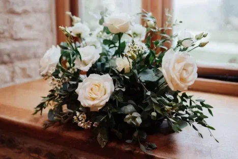 A bouquet of white roses and greenery on a wooden surface near a window.