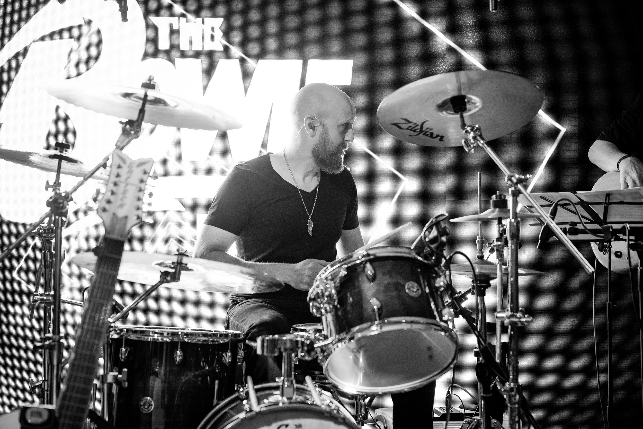 A bald bearded man playing drums on a stage, with cymbals and a large screen in the background displaying 'The Dive'.