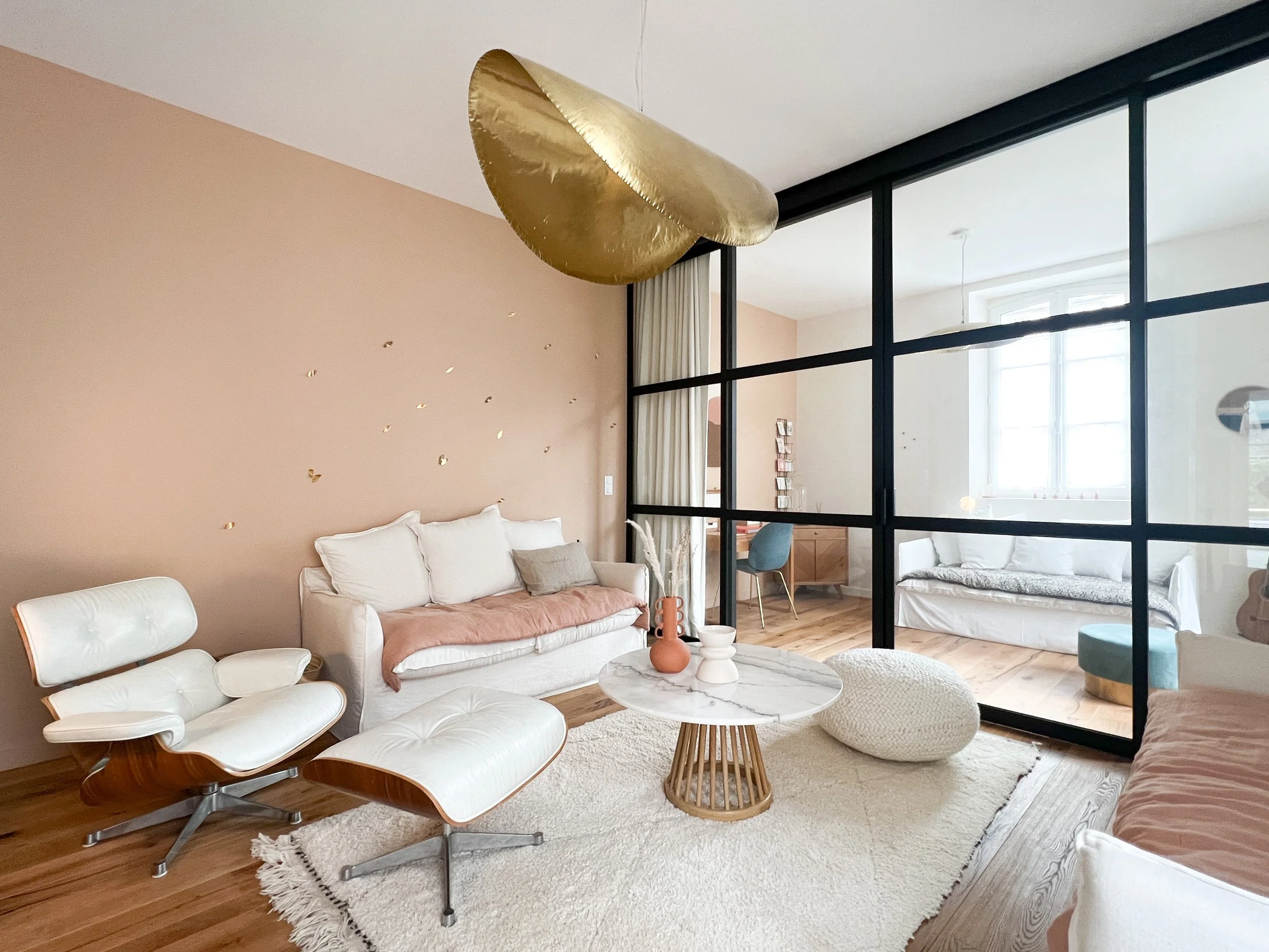 Rénovation d'un salon intérieur moderne avec canapé en lin blanc, fauteuil Eames designer en cuir blanc, table en marbre, mur beige décoré de pétales dorées,  verrière noir et pièce lumineuse avec fenêtres.