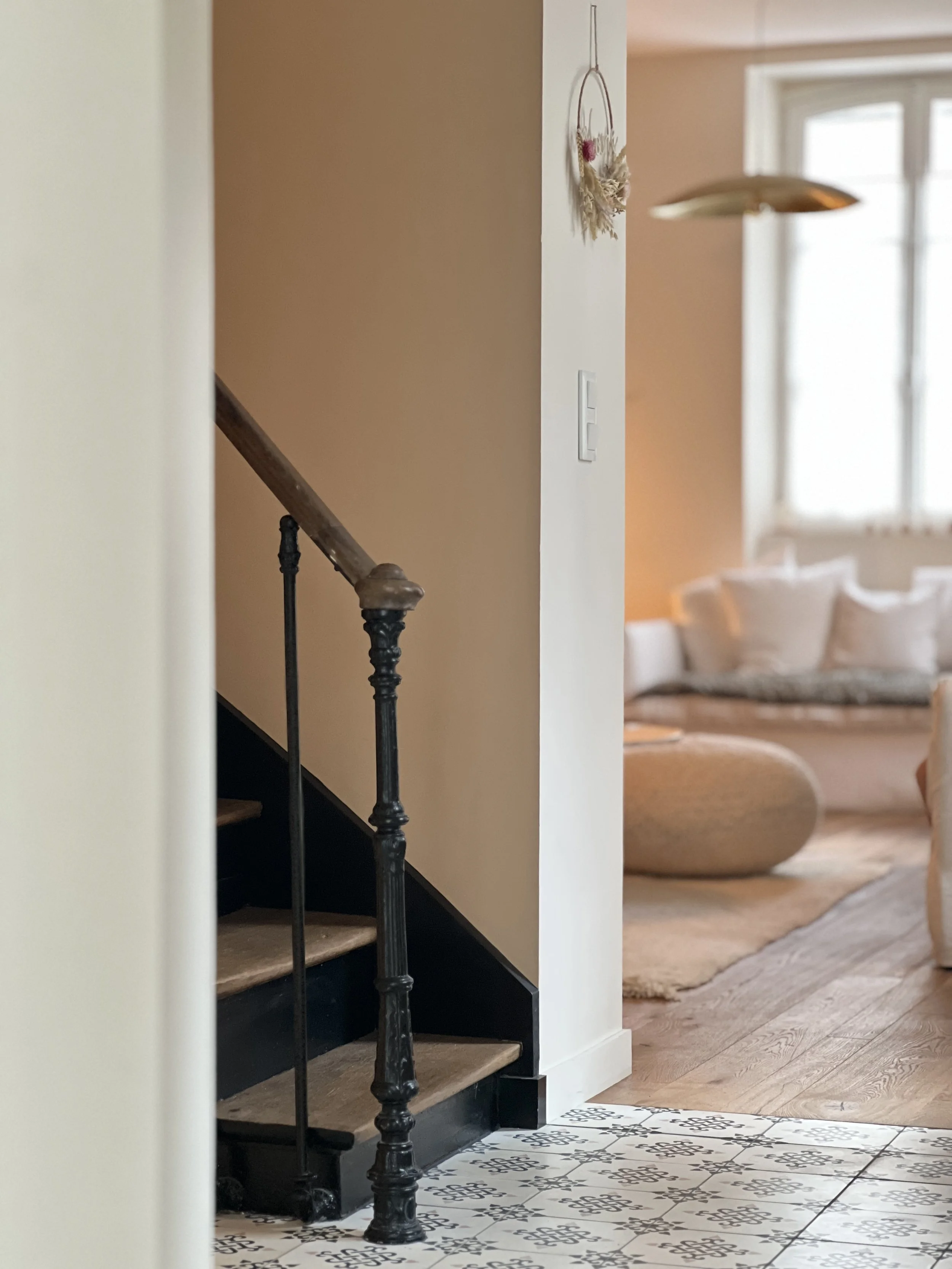 Escalier en bois avec une rampe en métal noir, sol en carreaux blancs avec motifs noirs, mur blanc, décoration murale suspendue avec fleurs séchées, pièce lumineuse avec canapé beige et tapis en laine, grande fenêtre laissant entrer la lumière naturelle.