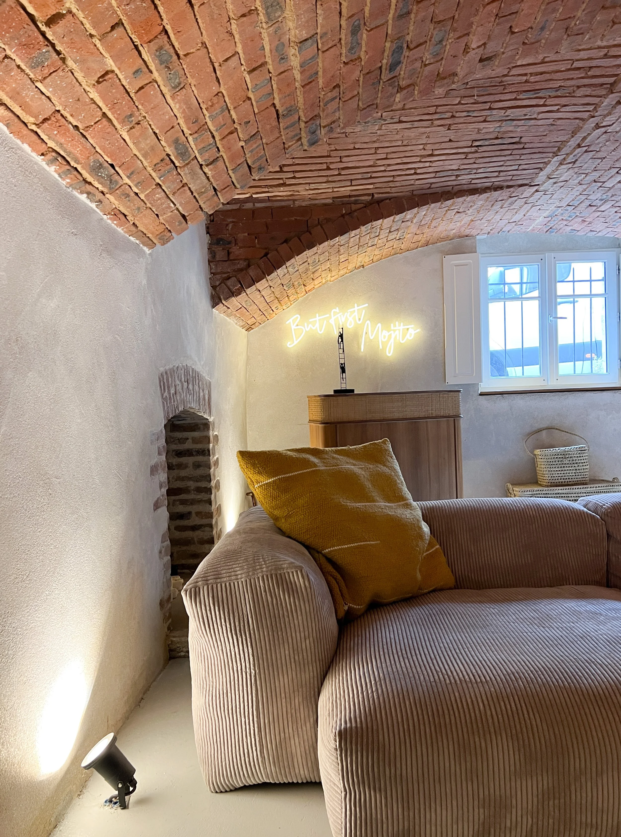 Une pièce avec un plafond voûté en briques, une fenêtre avec volets blancs, un canapé avec des coussins jaunes, une lampe de projecteur au sol et une décoration néon indiquant 'Bar Fast Mojito'.