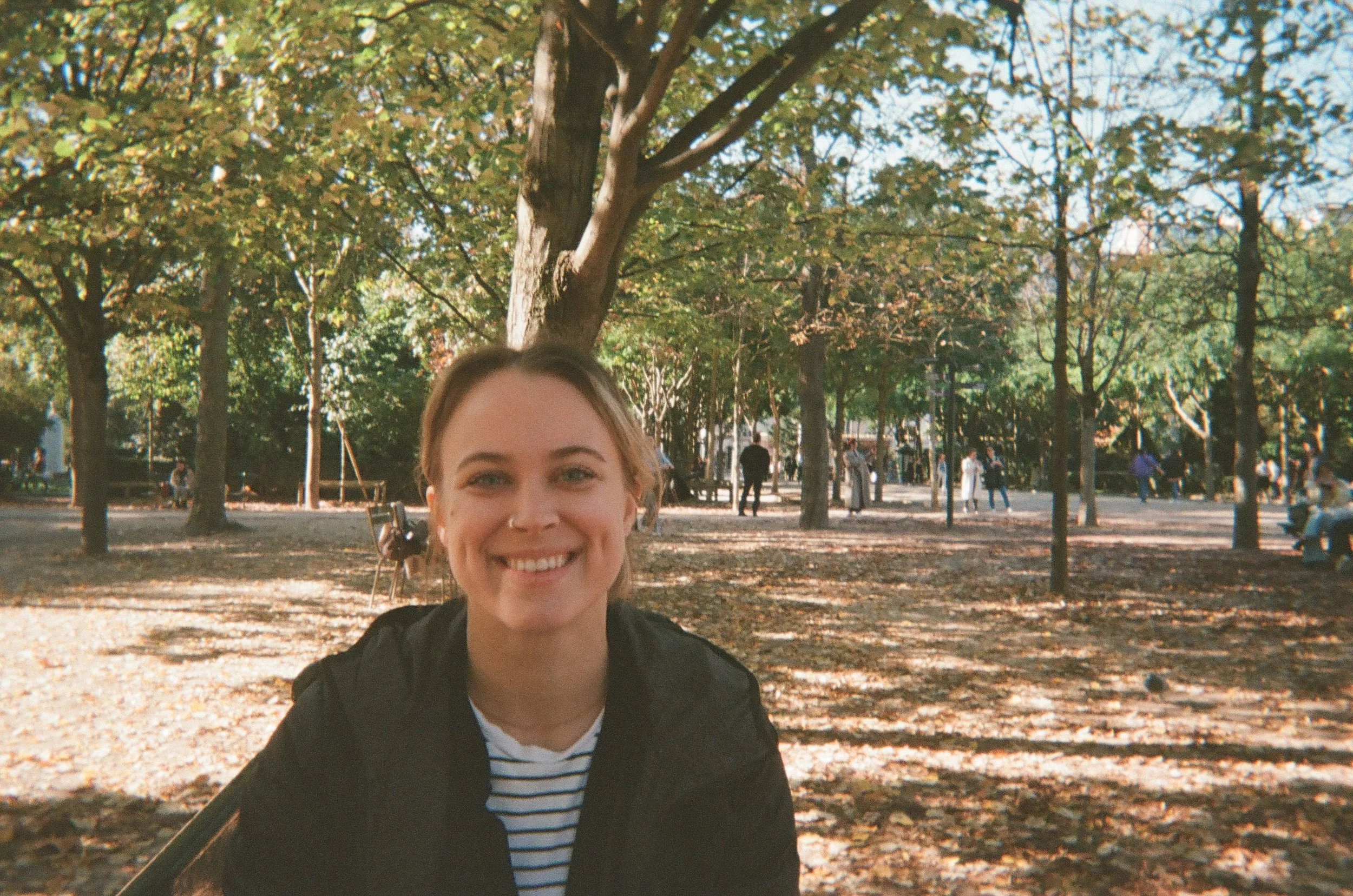 A young woman with blonde hair smiling at the camera in a park on a sunny day, surrounded by trees with green leaves and fallen autumn leaves on the ground, with people in the background.