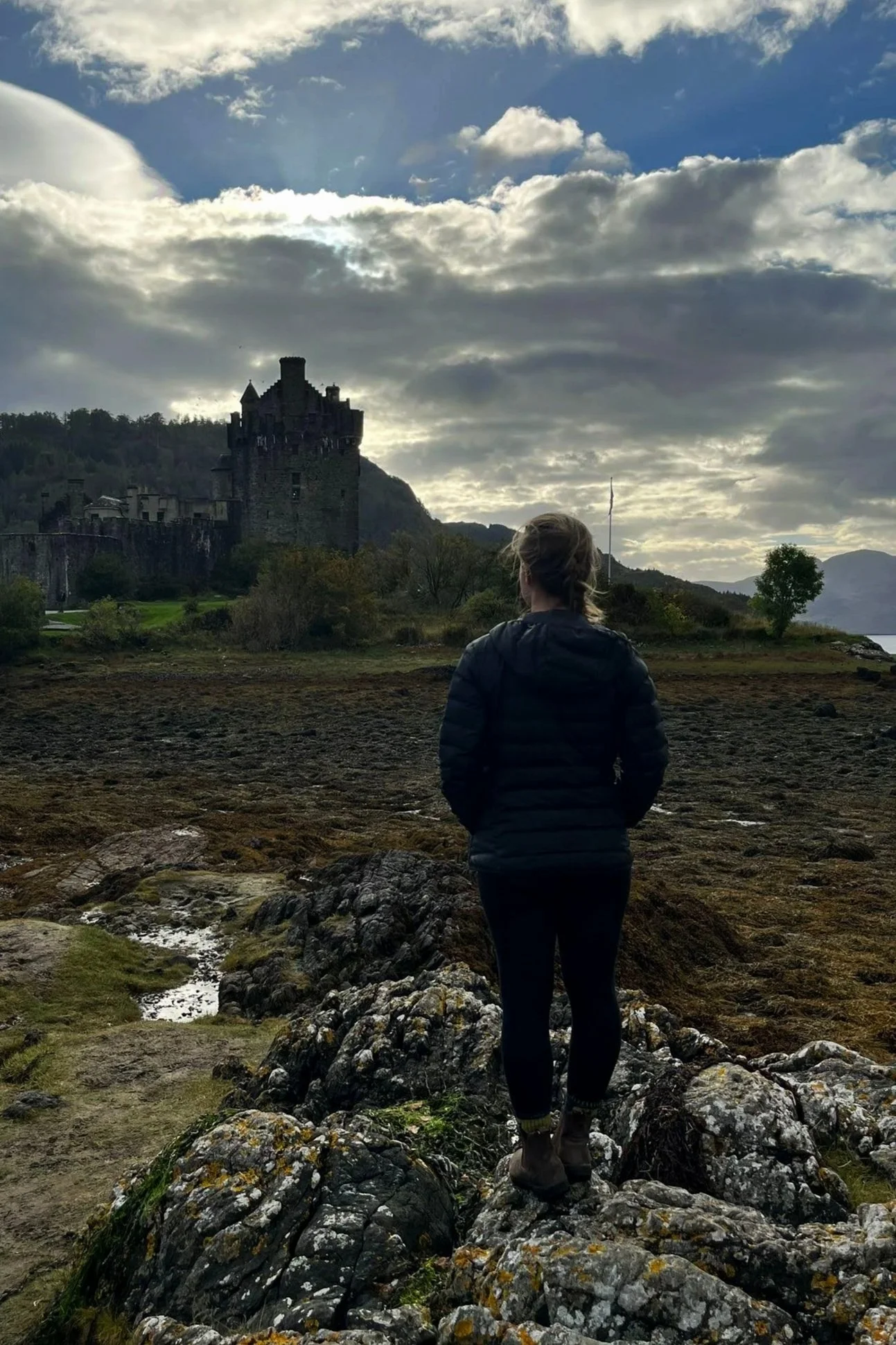 Megan's hair is in a braid, wearing a black puffer coat, black leggings, and brown boots. Eilean Donan Castle stands in background.