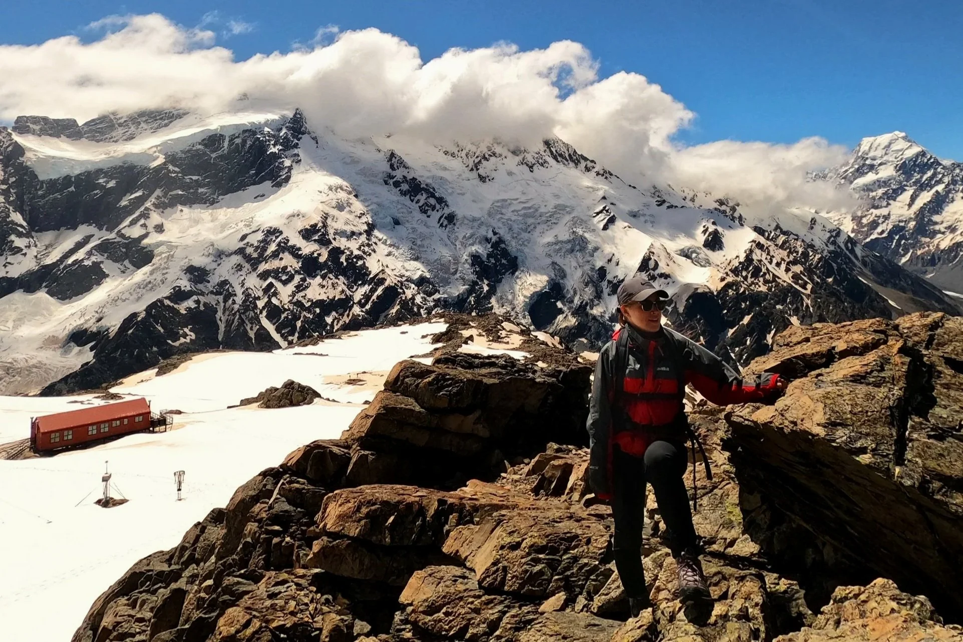 Megan standing in the foreground on a rocky terrain. in the background there is a red hut on a snowy plateau with snow covered mountain peaks behind.