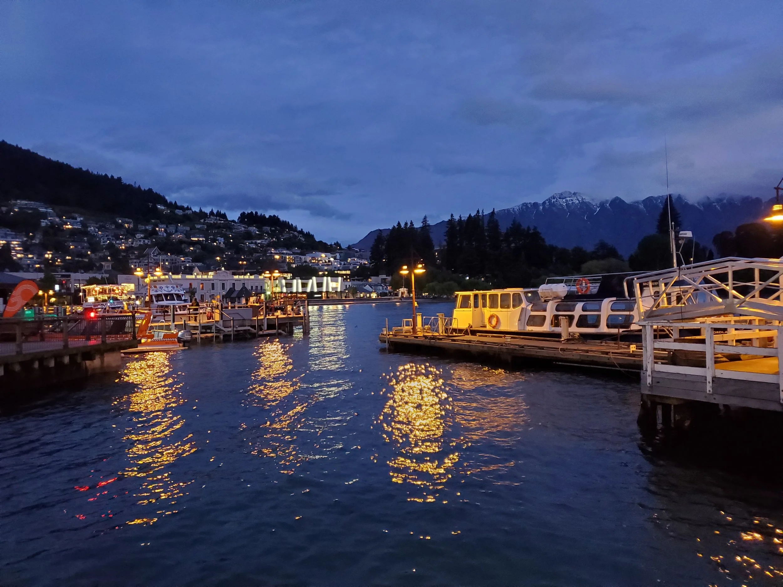 lights of Queenstown New Zealand reflecting on water at dusk. Mountains in background, boats in foreground