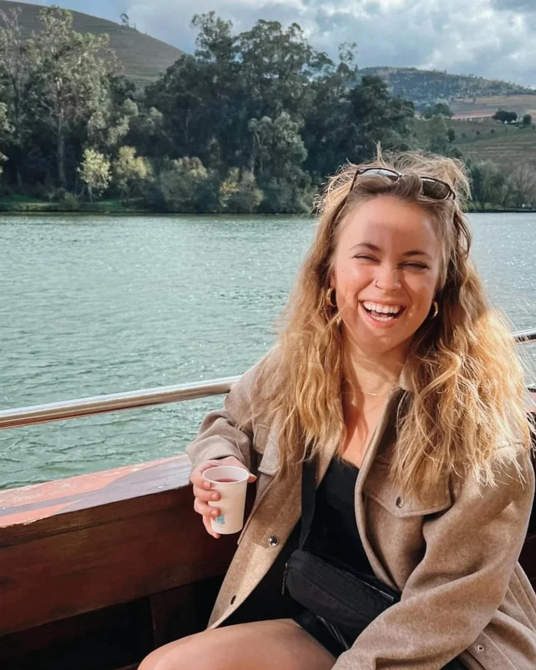 A woman with wavy blonde hair, wearing a tan jacket, sitting on a boat, holding a small cup, smiling and laughing by a river with green trees and hills in the background.