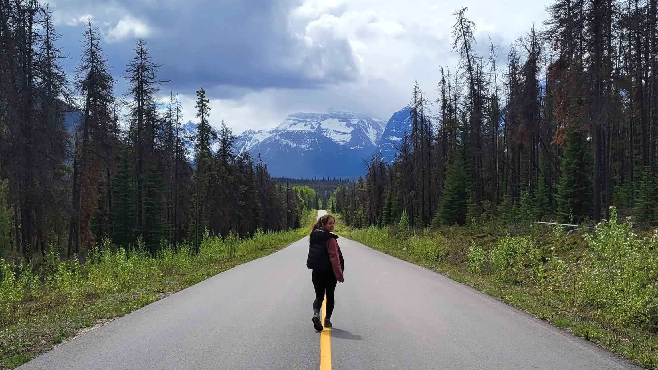 girl wearing a black vest and pink sweater walking down a road with no cars, pine trees line the road, moutains are in the distance, she is looking back over her shoulder at the viewer