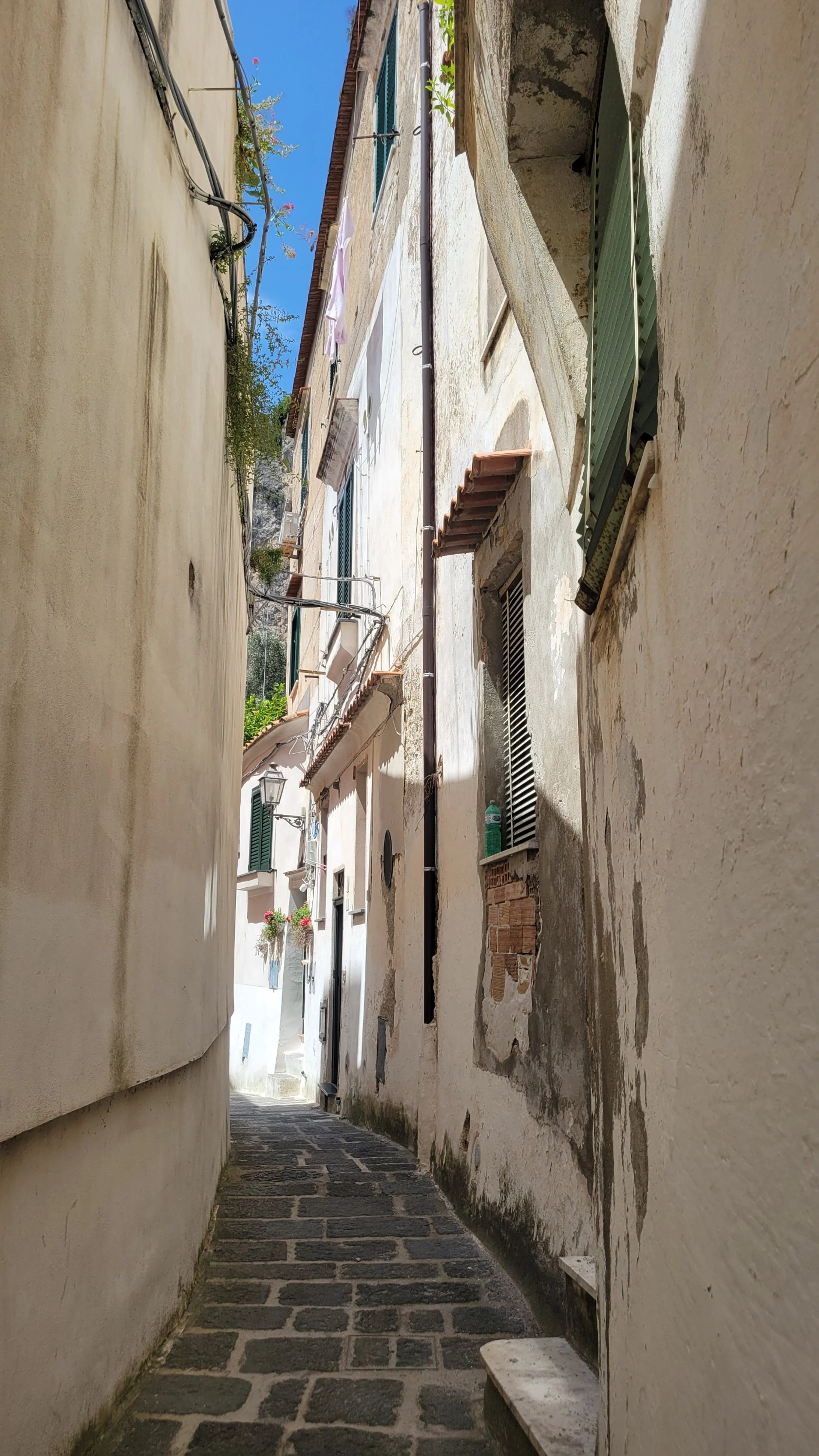 white houses line narrow pedestrian path made of cobblestones. a green awning, terracotta shingles and a narrow opening to blue skies above
