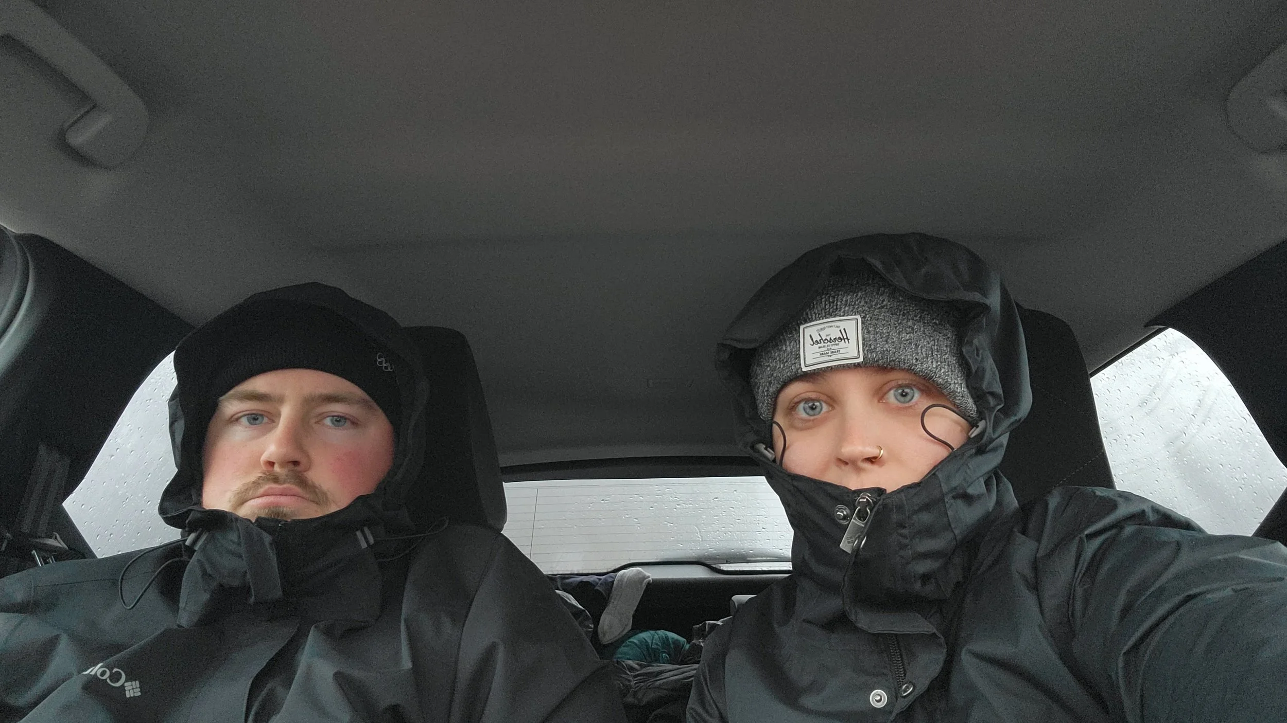 Two people sitting in the back of a vehicle, wearing black weatherproof jackets and knit hats, with rain visible through the windows.
