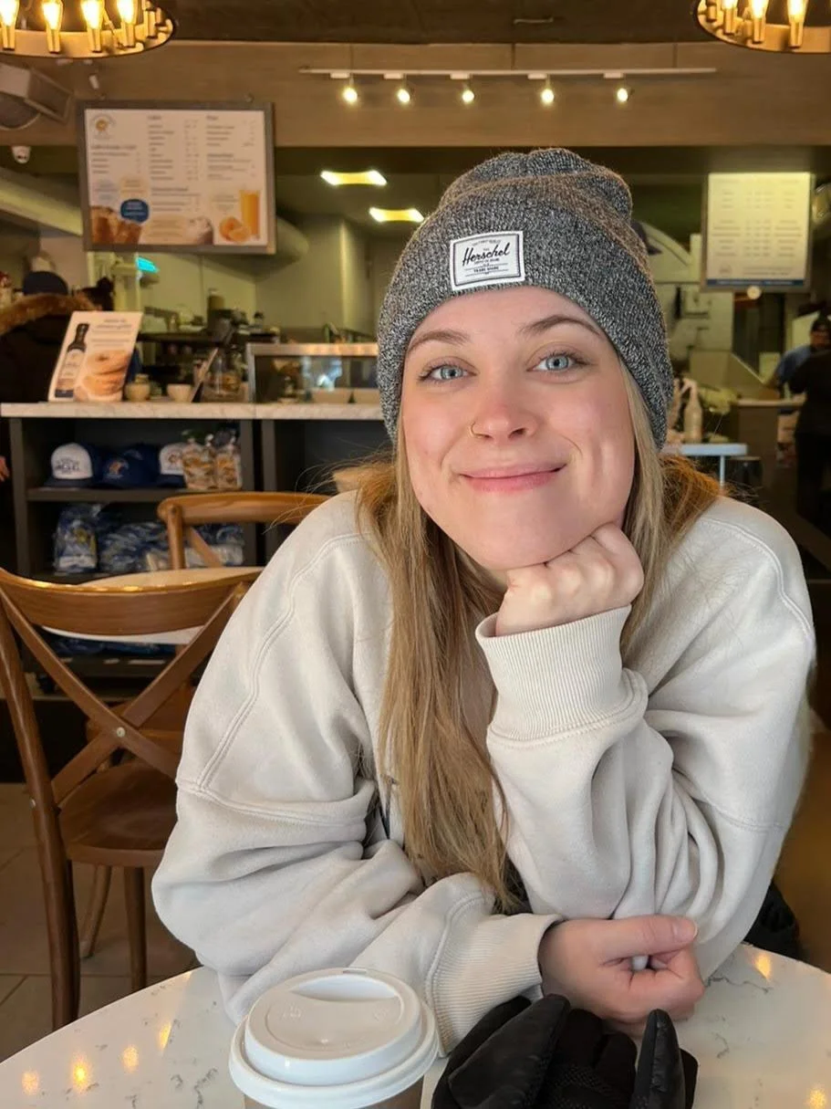 wearing a grey toque and white sweater, smiling at the camera. At a table waiting for st viateur bagel in Montreal