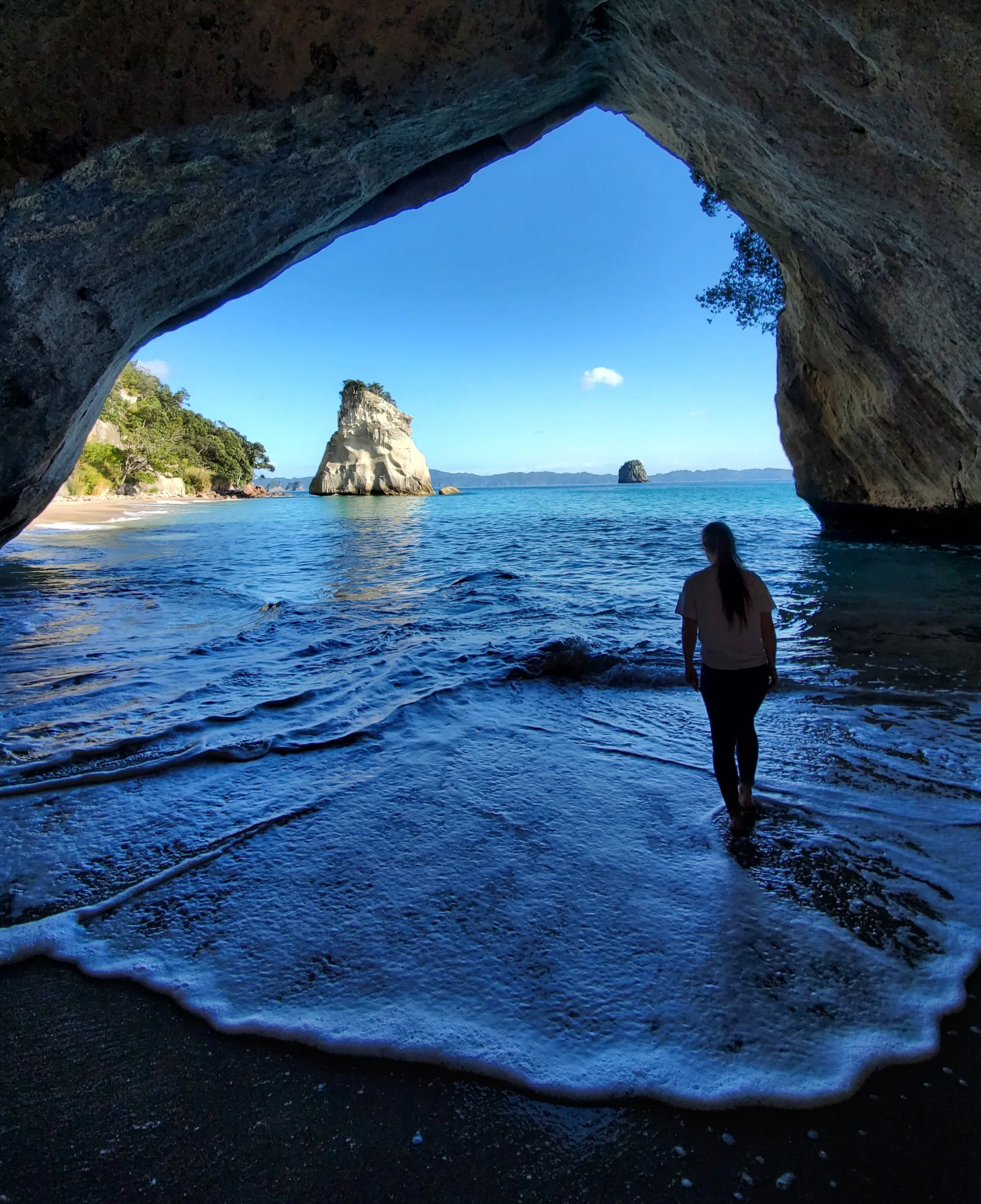 megan in a white tee and jeans walking away into the sea foam. The cave opens up to a view with blue ocean and sky and a prominent white rock jutting out of the water.