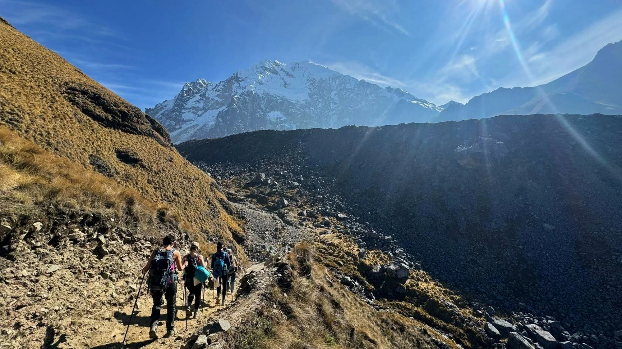 Four hikers with poles and day packs walking away from the camera. In the background are snow capped mountains and blue skies.
