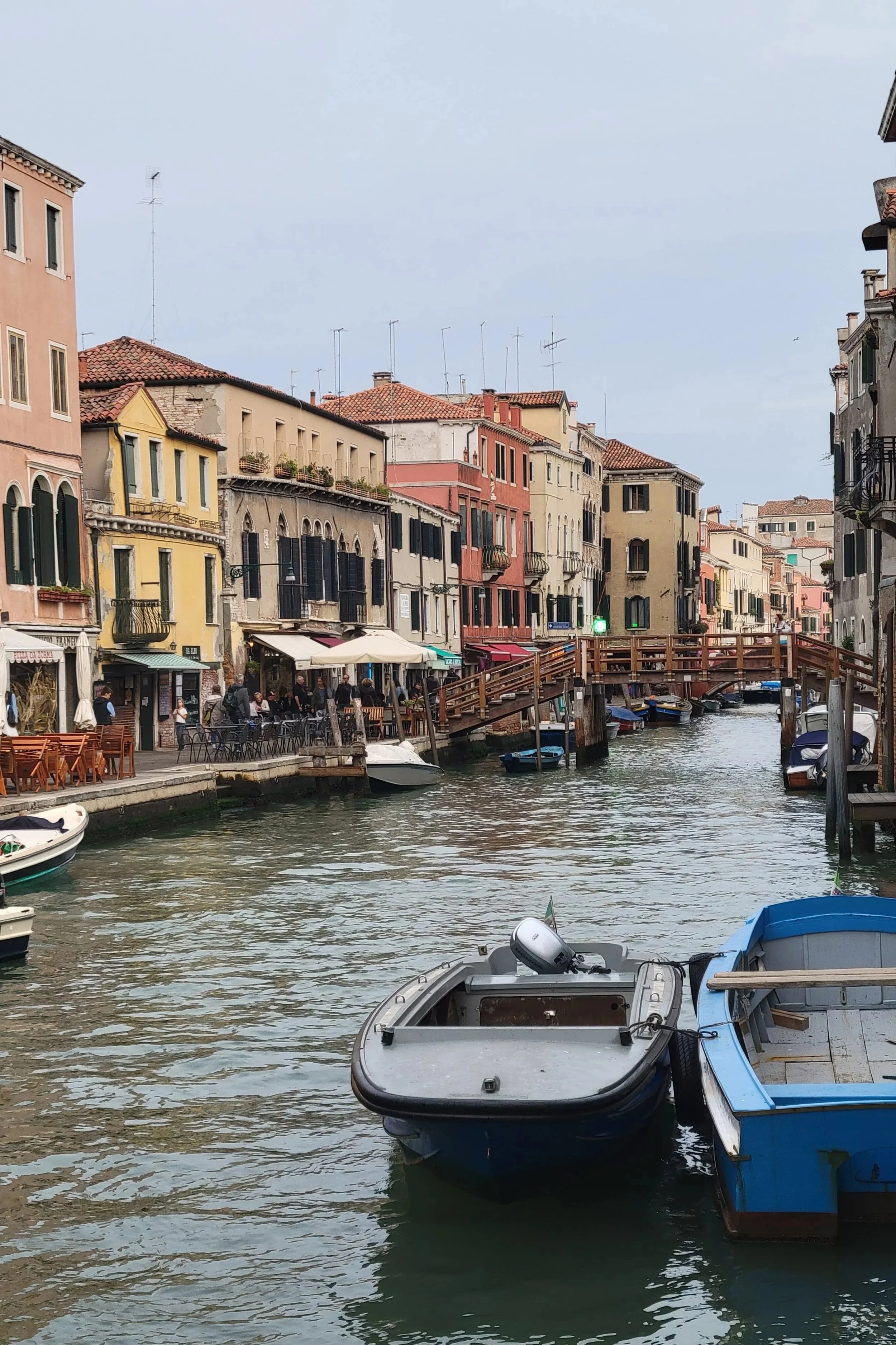 many boats tied to the edge of canal, colourful, old buildings line the sidewalk. a wooden pedestrian bridge crosses the canal