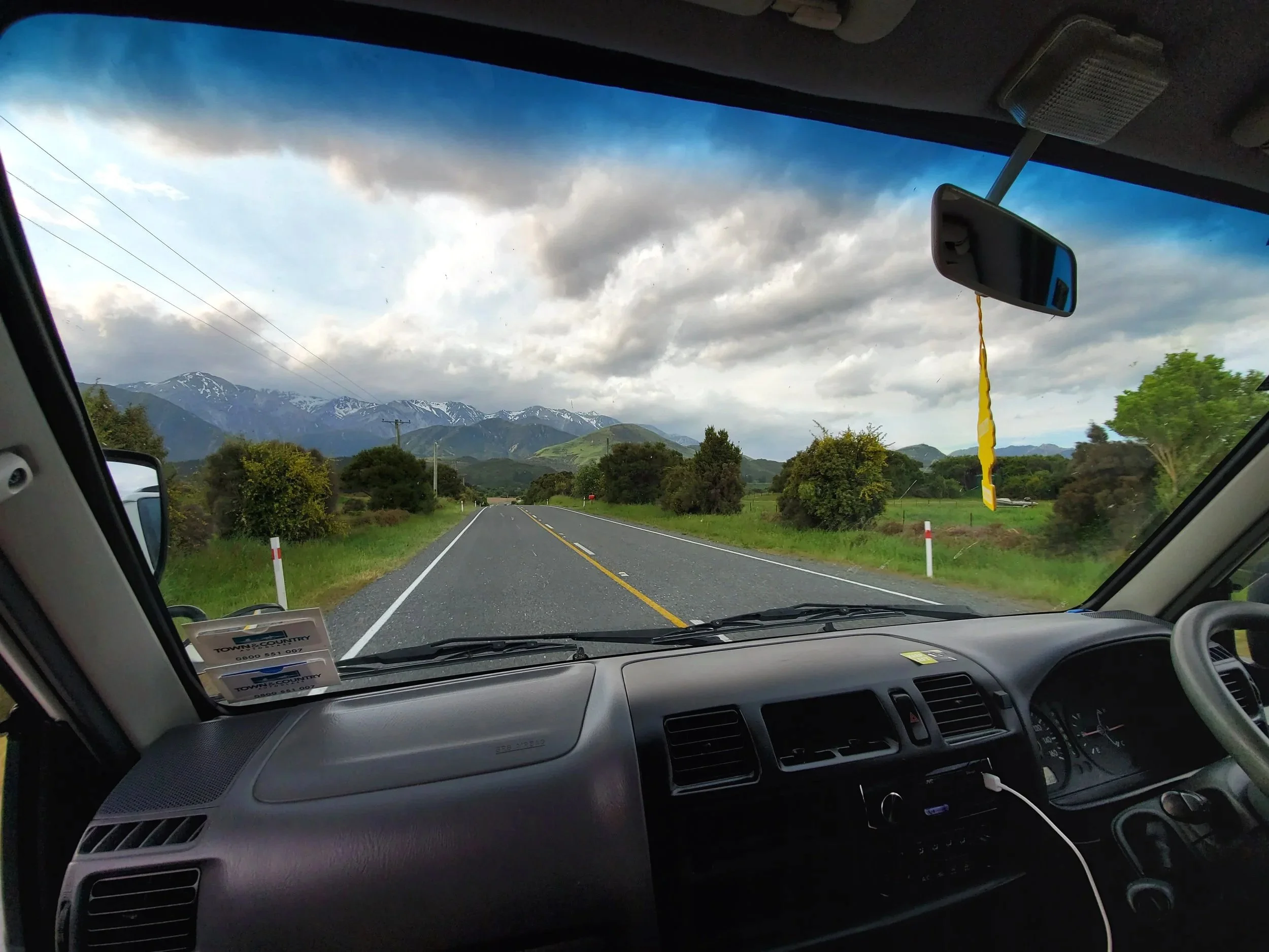 Open road with mountains in background seen through dashboard of campervan. Yellow air freshener hangs from the rearview mirror