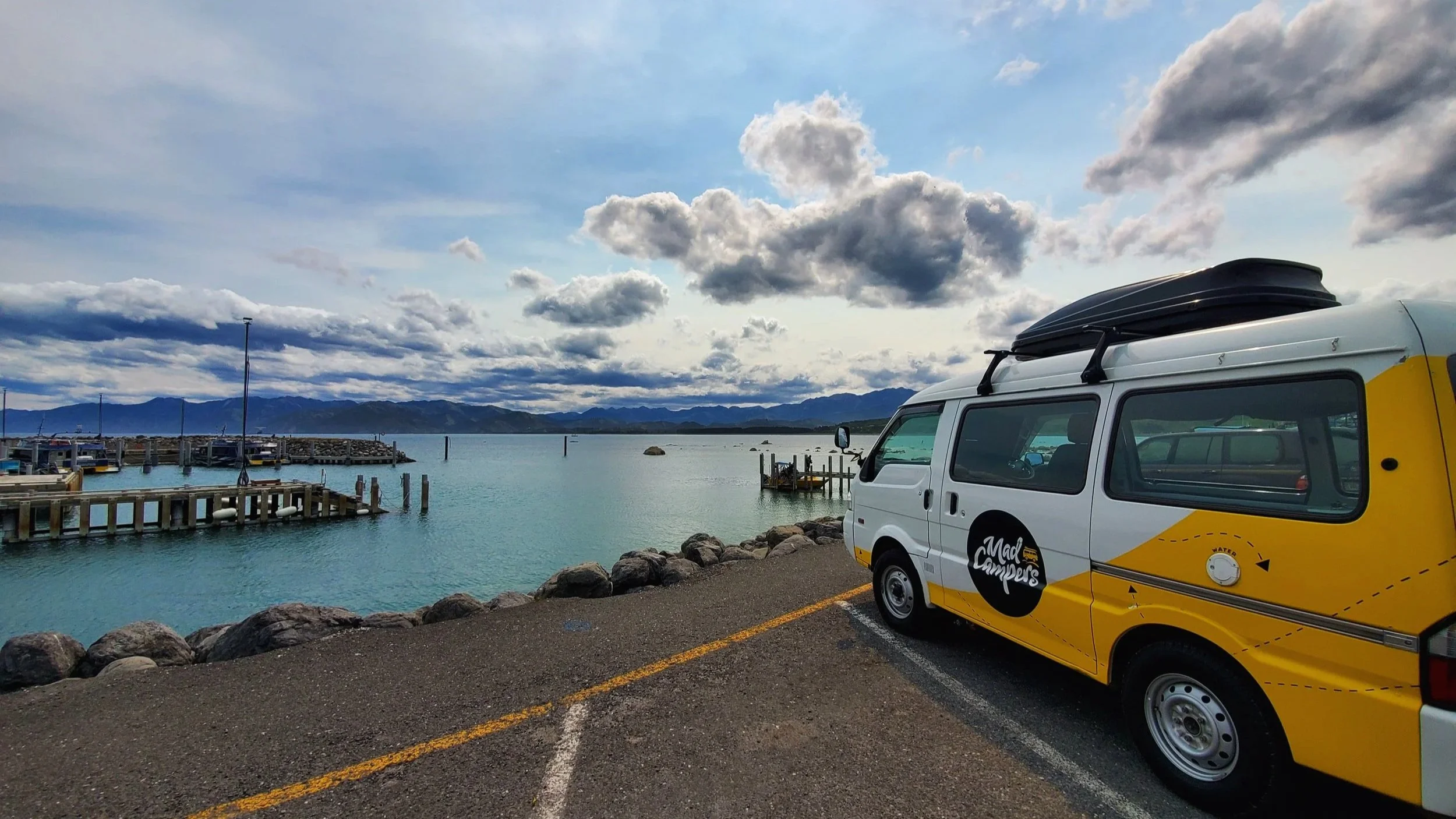 Yellow and white campervan with "Mad Campers" logo parked beside ocean water, mountains in the background