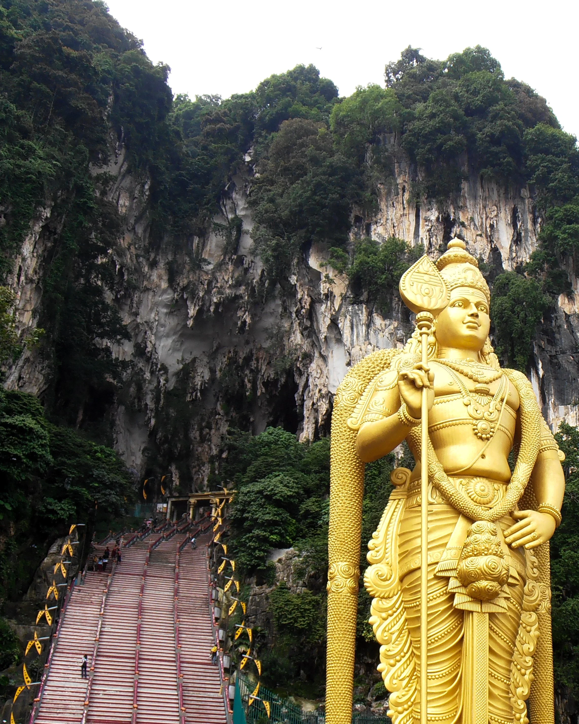 The entry to the Batu Caves temple seen at the top of a long red staircase. In the foreground there is a giant golden statue of Murugan.