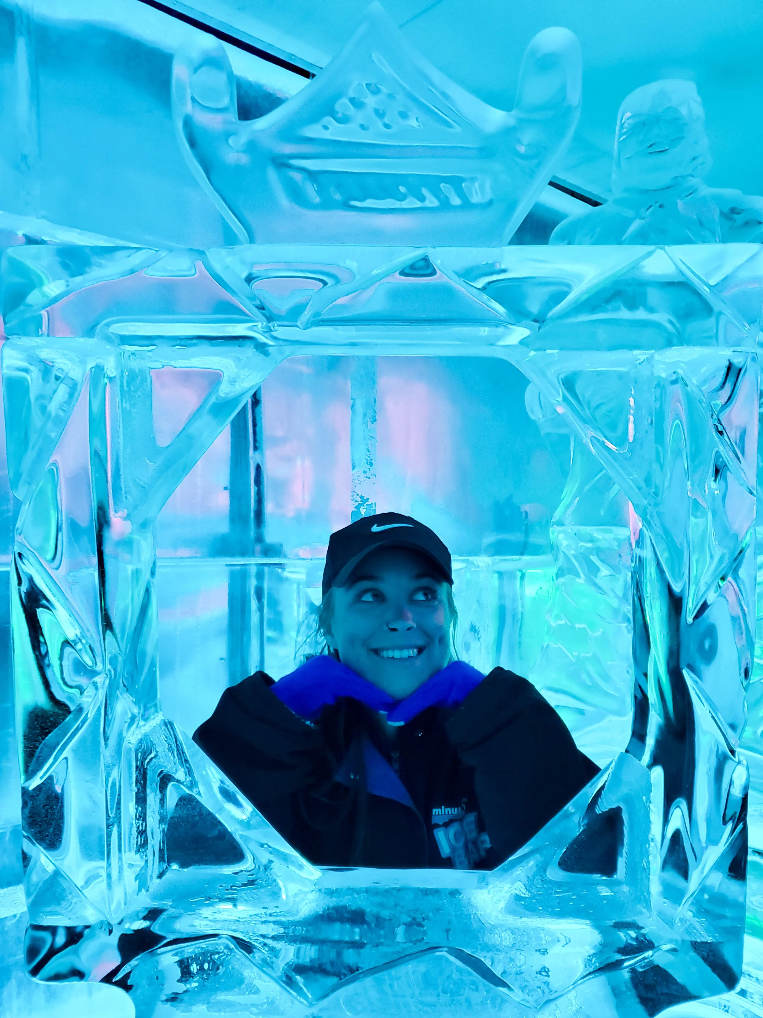 Me posing, framed by an ice sculpture. There is a cool blue tint to the lighting. I am wearing a black nike cap, purple gloves and a winter jacket.