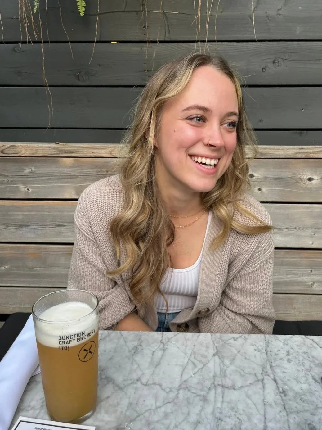 A young woman with long, wavy blonde hair, smiling and looking to her left, sitting at a table with a glass of beer labeled 'Junction Craft Brewery' in front of her, in an outdoor setting with a wooden fence background.