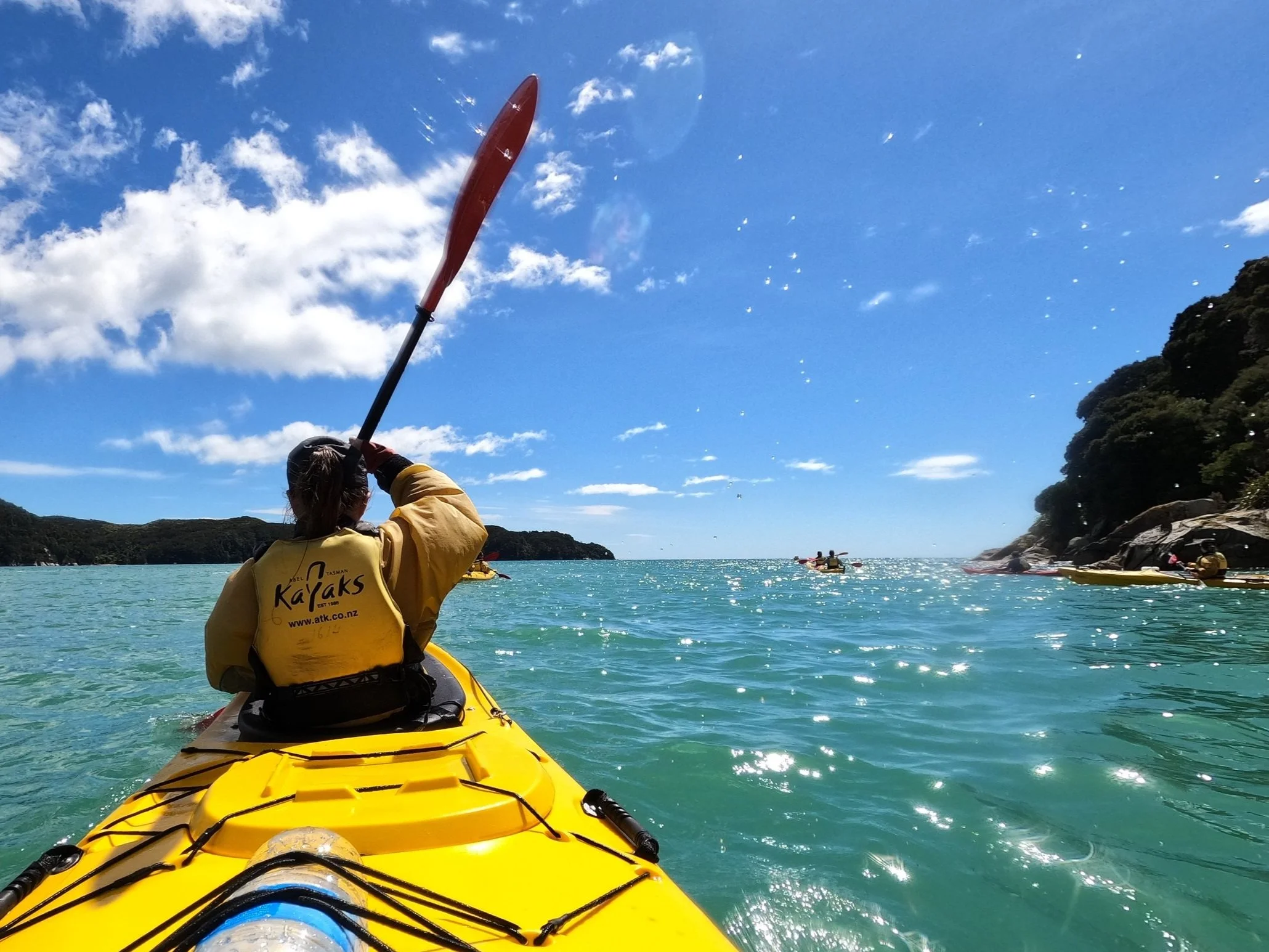 Megan wearing yellow life jacket in a yellow kayak paddling in the blue ocean water. Blue skies with a few clouds, and tree covered coasts