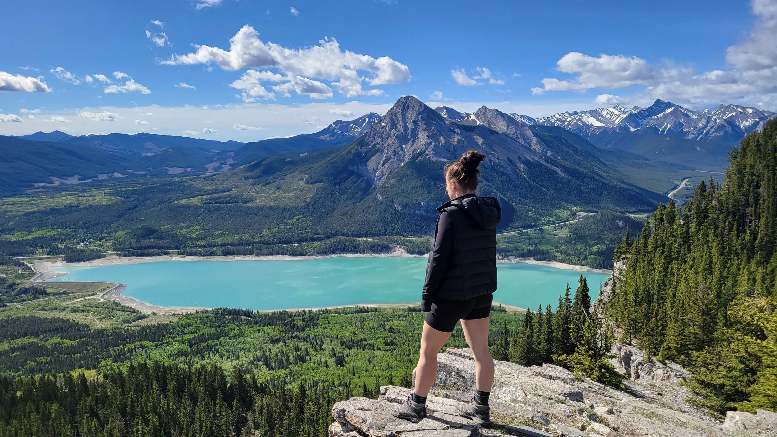 girl standing on a rocky shelf overlooking the forest below with a bright blue lake and mountains in the distance