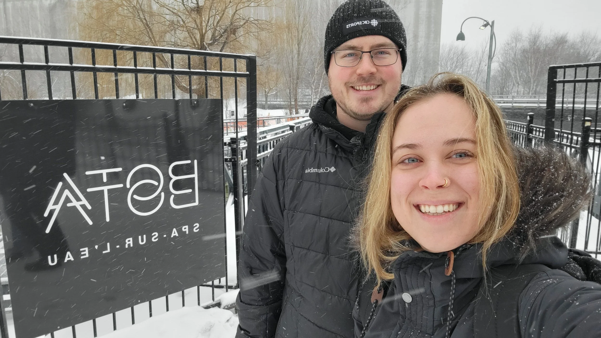 Tyson and Meg smiling at the camera bundled in winter layers. Standing beside bota bota sign in montreal