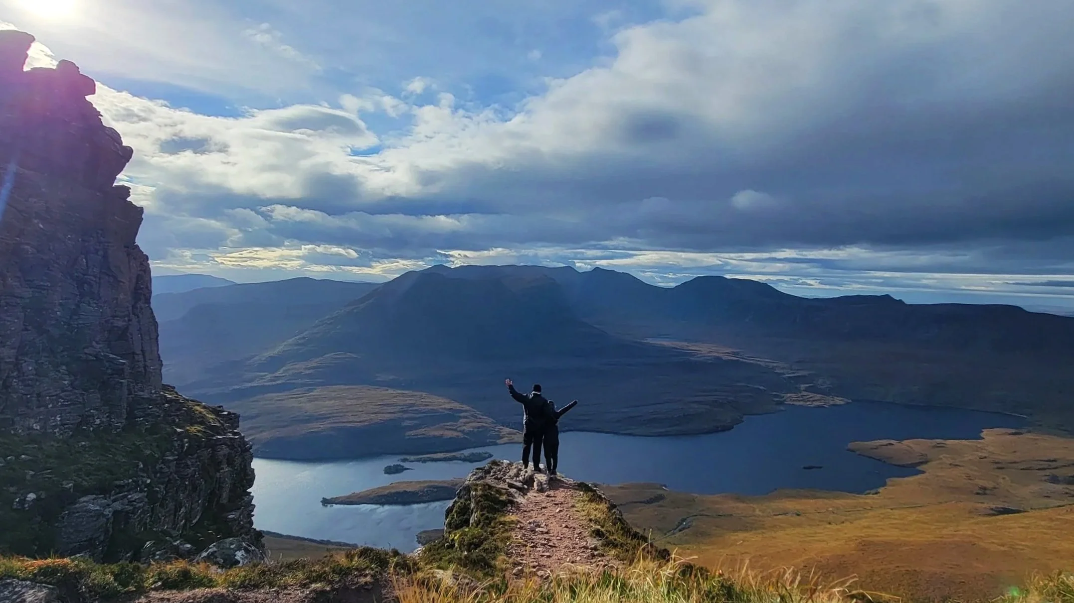 Megan and Tyson at top of Stac Pollaidh sun peaks through layer of clouds loch below mountains in background