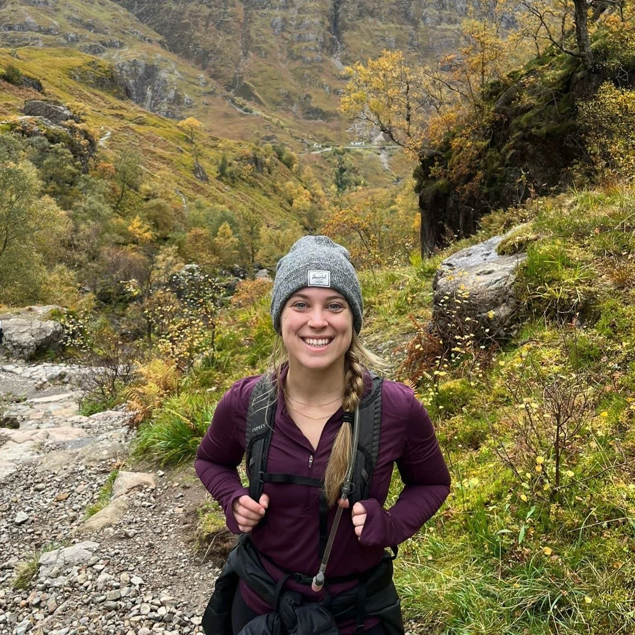 A smiling woman with a gray knit beanie and a purple jacket hiking on a rocky trail in a lush, autumn landscape with trees and mountains.