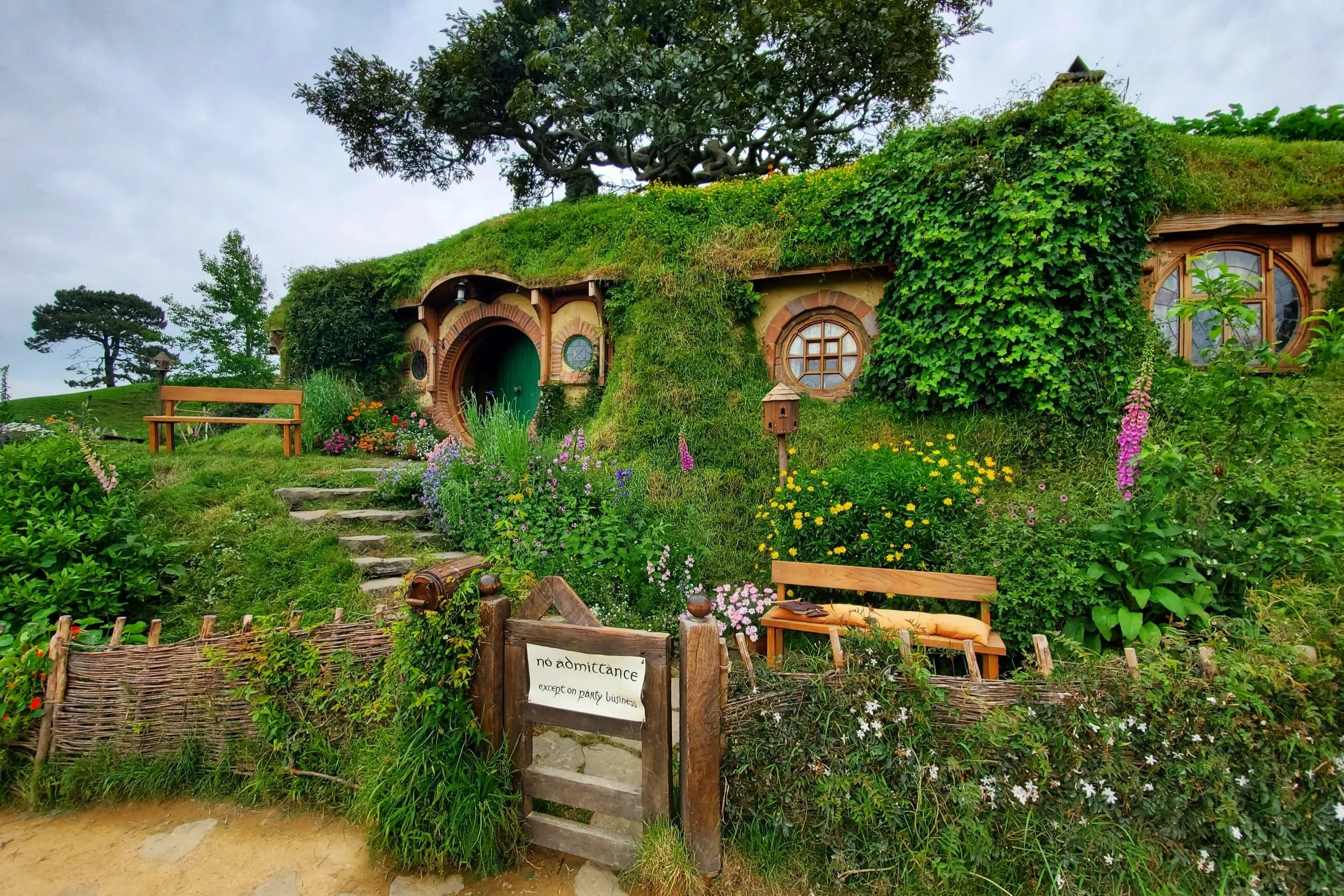 hobbit house built into the hill, grass grows on its roof. a gate infront of the house says "no admittance except on party business". colourful flowers grow in the garden. The front circular door is painted green