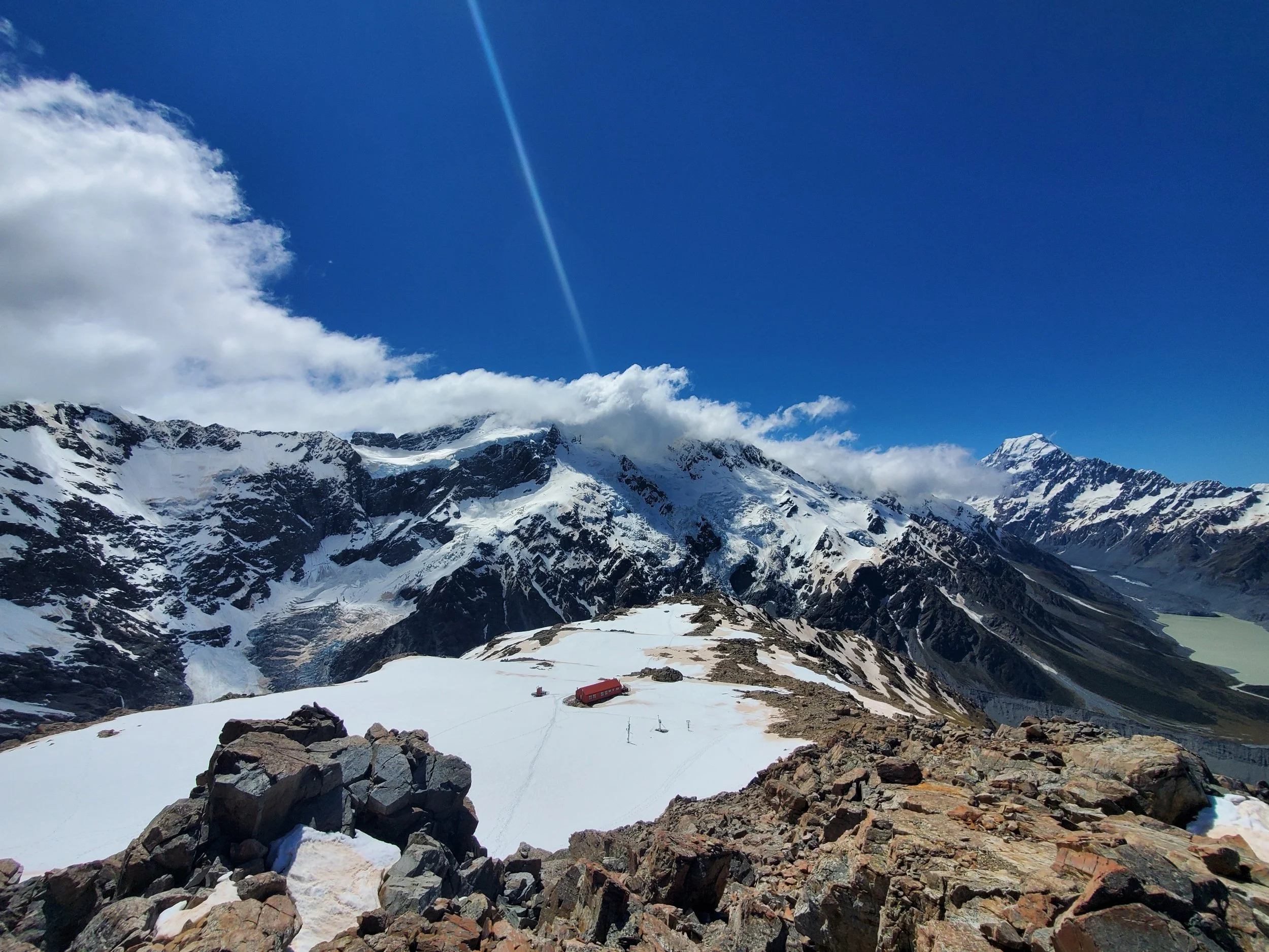 Red roof of Mueller Hut sitting on the snow covered peak. Large boulders perched along the edge, view of Hooker Lake in the Valley below with Mount Cook and other mountains in the background.