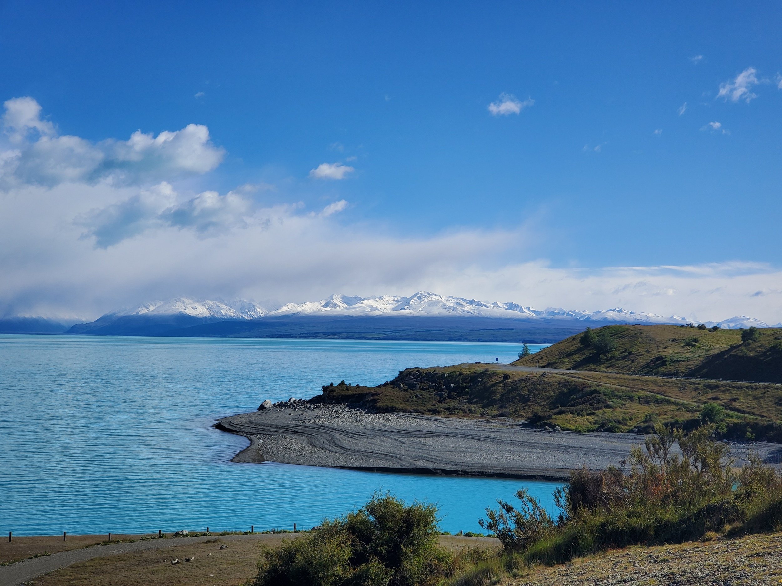 turquoise Lake Pukaki with snowy mountain range behind. Alps 2 Ocean trail winds along the coast.