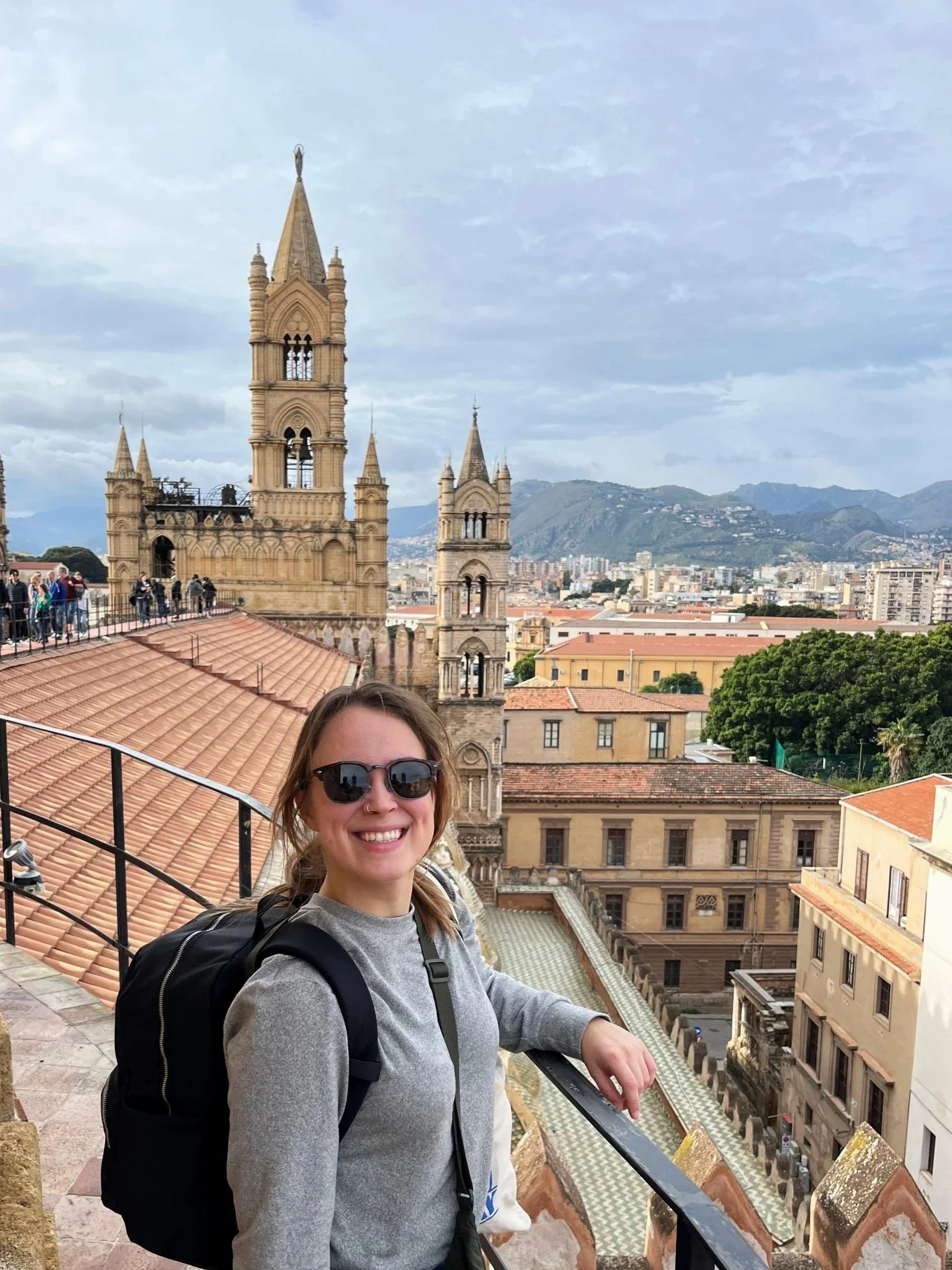 Girl smiling wearing sunglasses, black backpack and grey longsleeve standing on the roof of the Palermo Cathedral. In the background is the city and a mountain outline