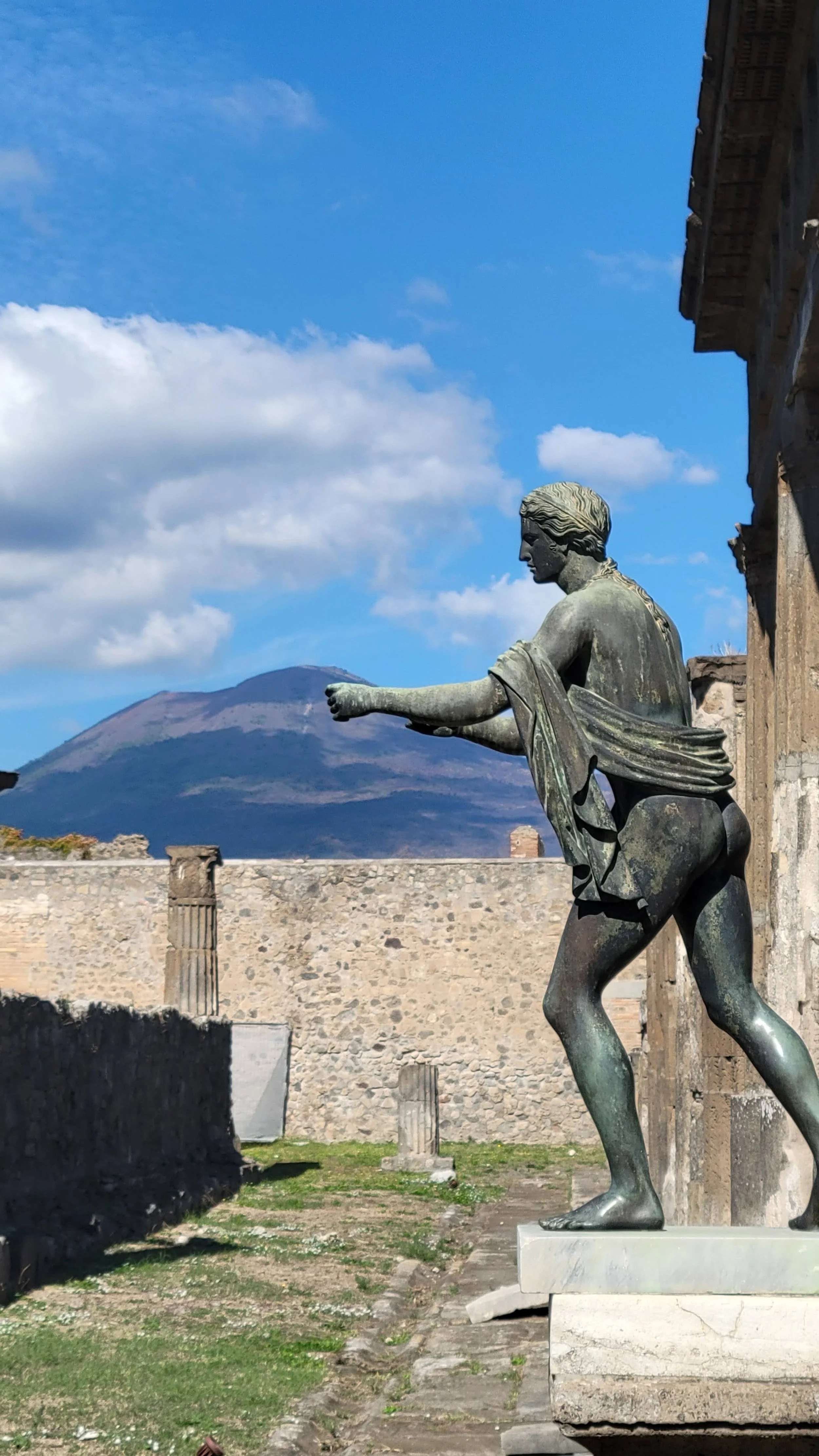 A bronze statue standing tall, remnants of columns and walls. Vesuvius stands in the background