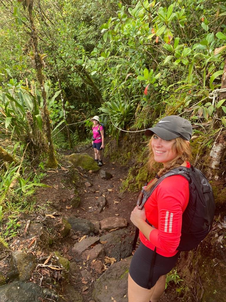 Megan smiling at camera wearing black cap, pink top, black biker shorts, black day pack. Heading down the trail through lush rainforest