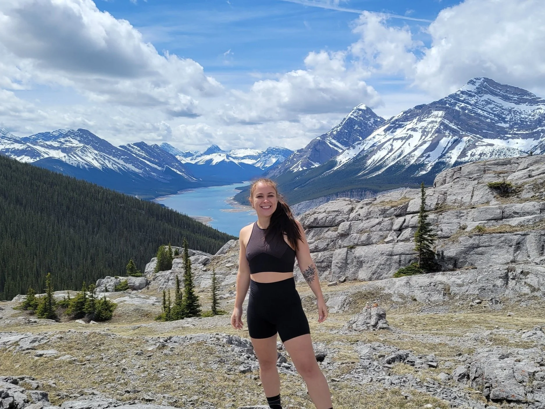 A woman smiling and standing in a mountainous landscape with snow-capped peaks, a lake, and a partly cloudy sky in the background.