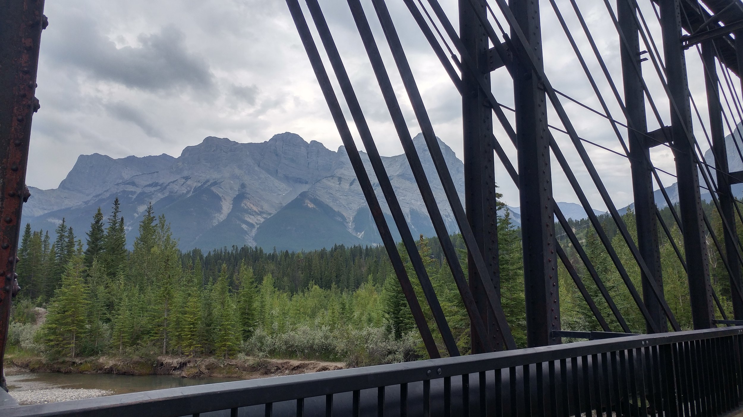 view of the mountains in the background with trees lining the bow river in canmore. The photo is taken from the old railway bridge, its steel structure is in the foreground