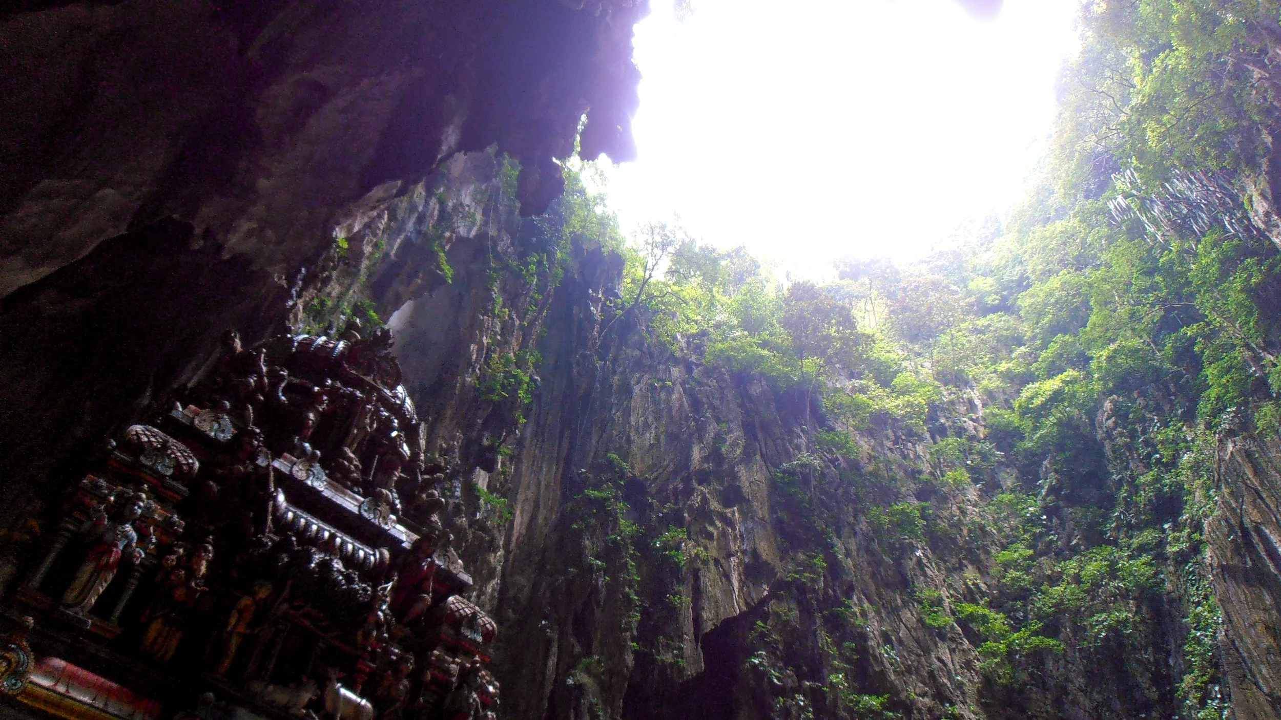 Inside the Batu Caves, looking up toward the opening in the roof of the cave as light flows in. The top of the temple can be seen in the bottom left corner.