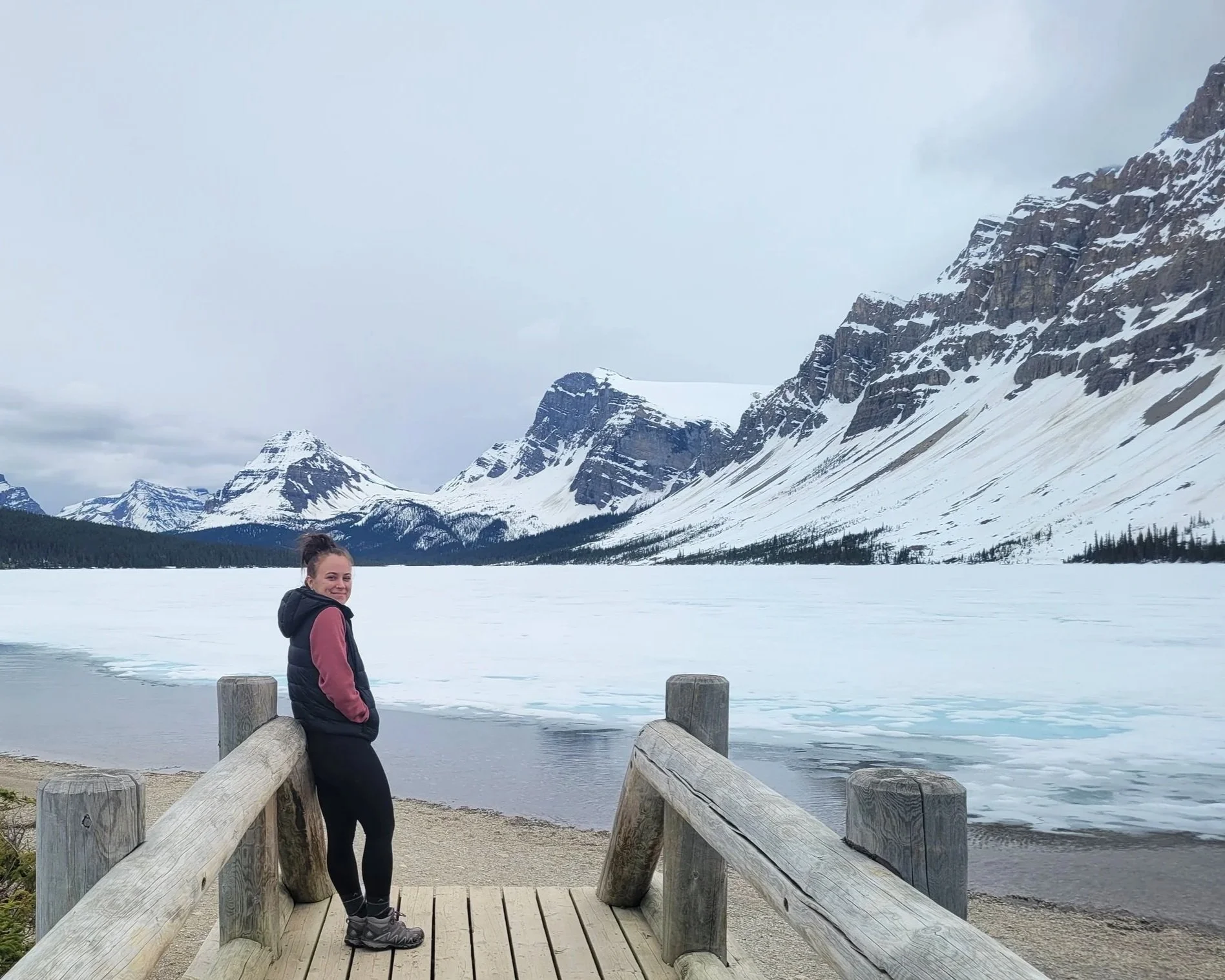 girl smiling at camera wearing black vest, pink sweater, black leggings, hiking boots. A frozen lake behind her with glacier covered mountains in the distance.