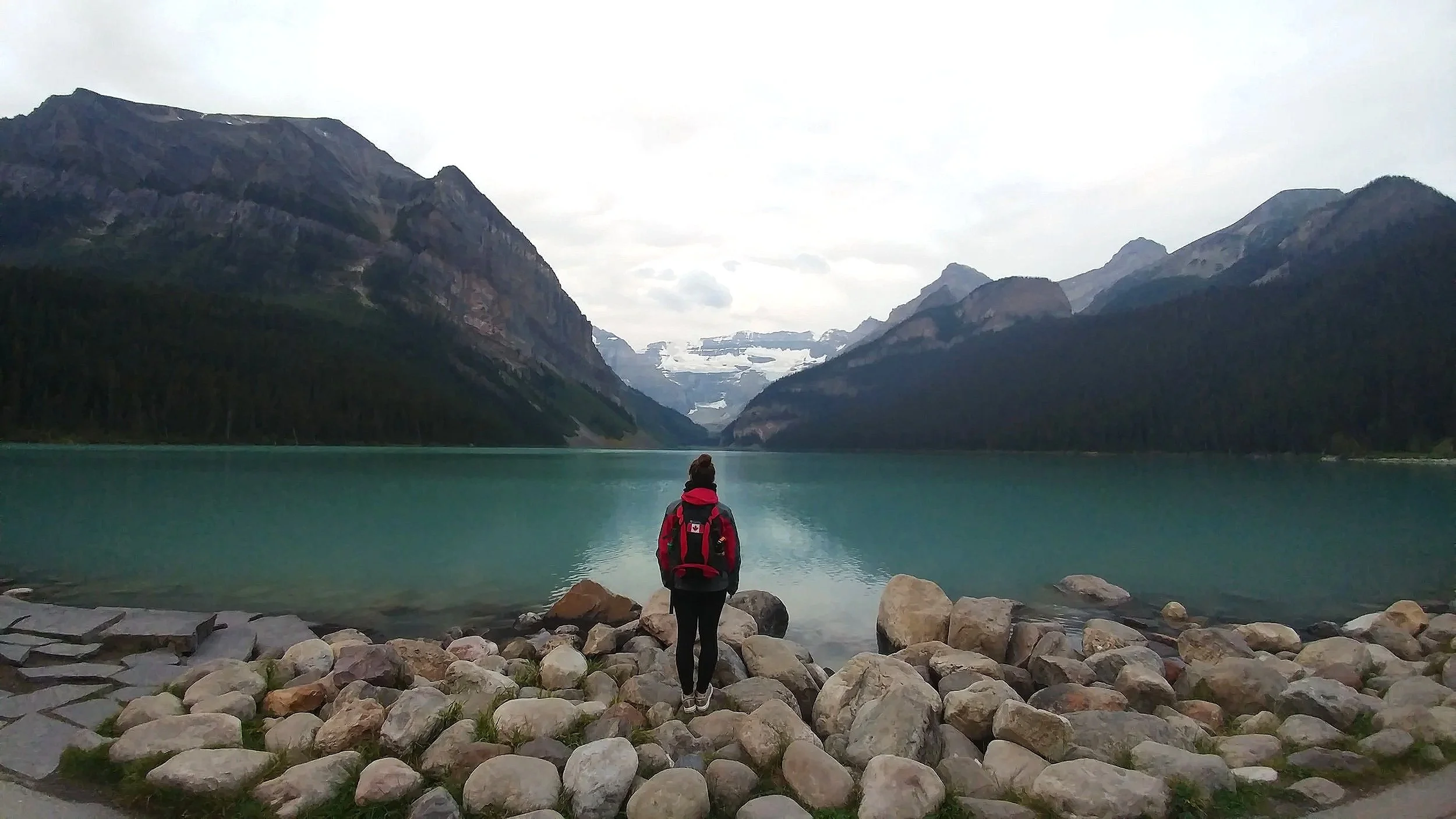 girl looking out over a green-blue lake with mountains and a glacier in the distance. She is wearing a red jacket and a red and black backpack with a canadian flag on it.