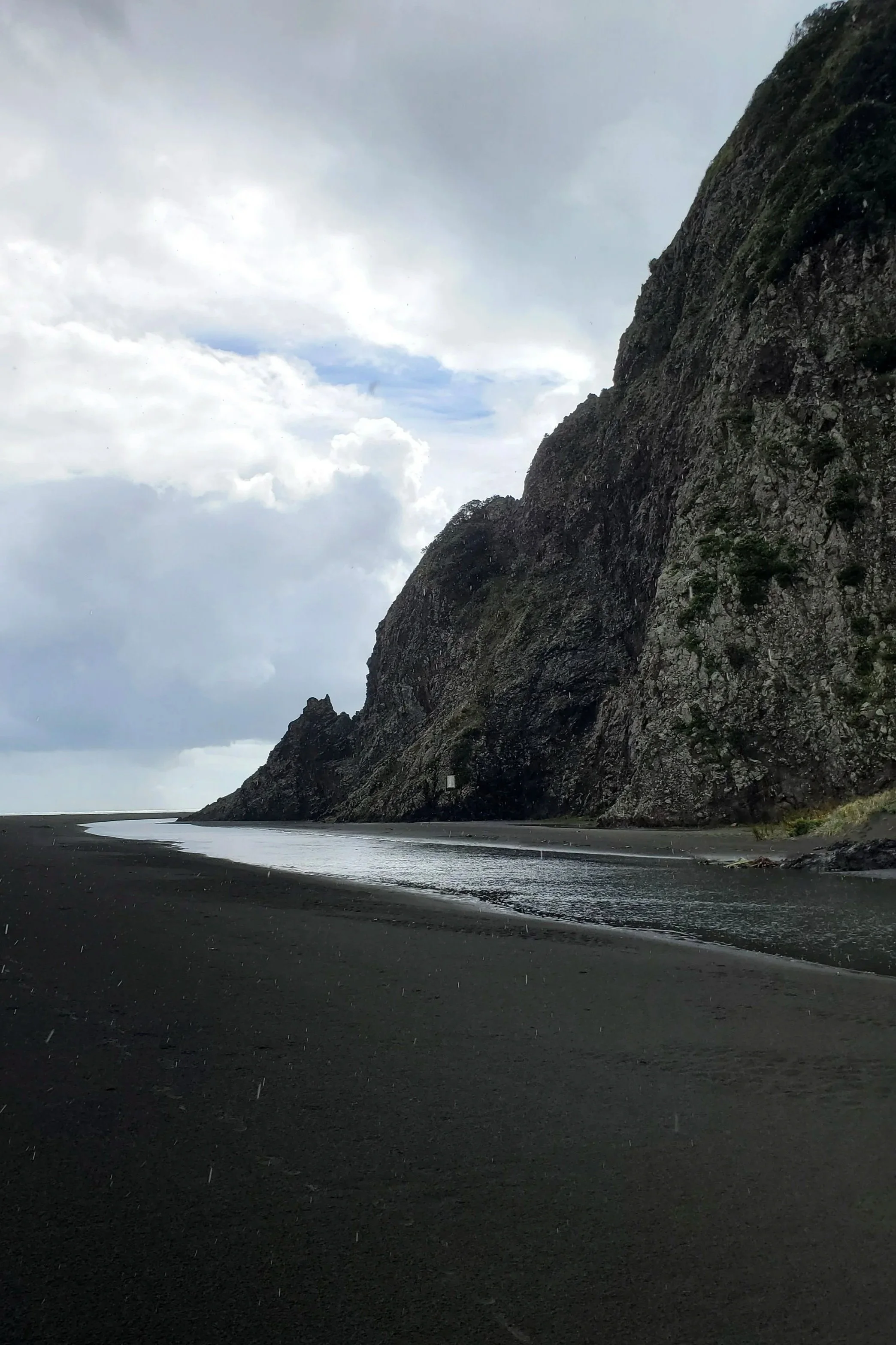 black sand beach a narrow stream of water heading for the ocean through the sand black cliffs just up into the sky. lightly raining