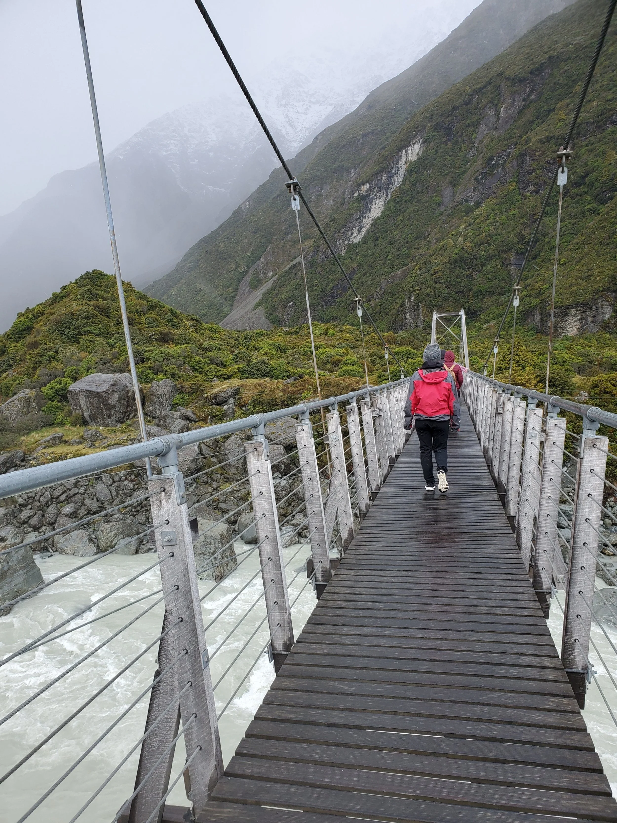 Megan wearing a red raincoat and grey hat, walking across a suspension bridge over a river through a valley lined by towering mountains