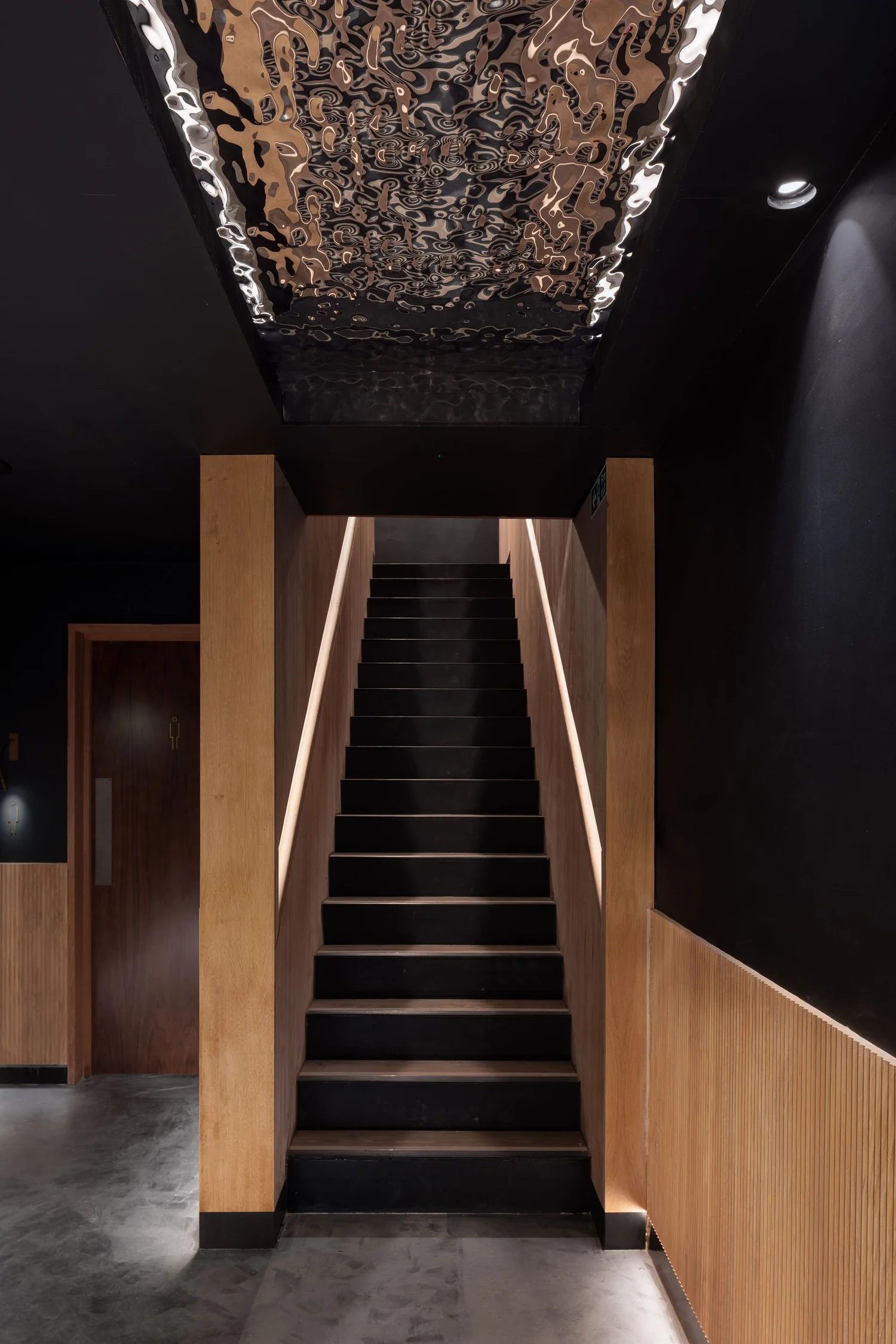 Timber-lined staircase at Soom with illuminated handrails beneath a rippled reflective ceiling panel.