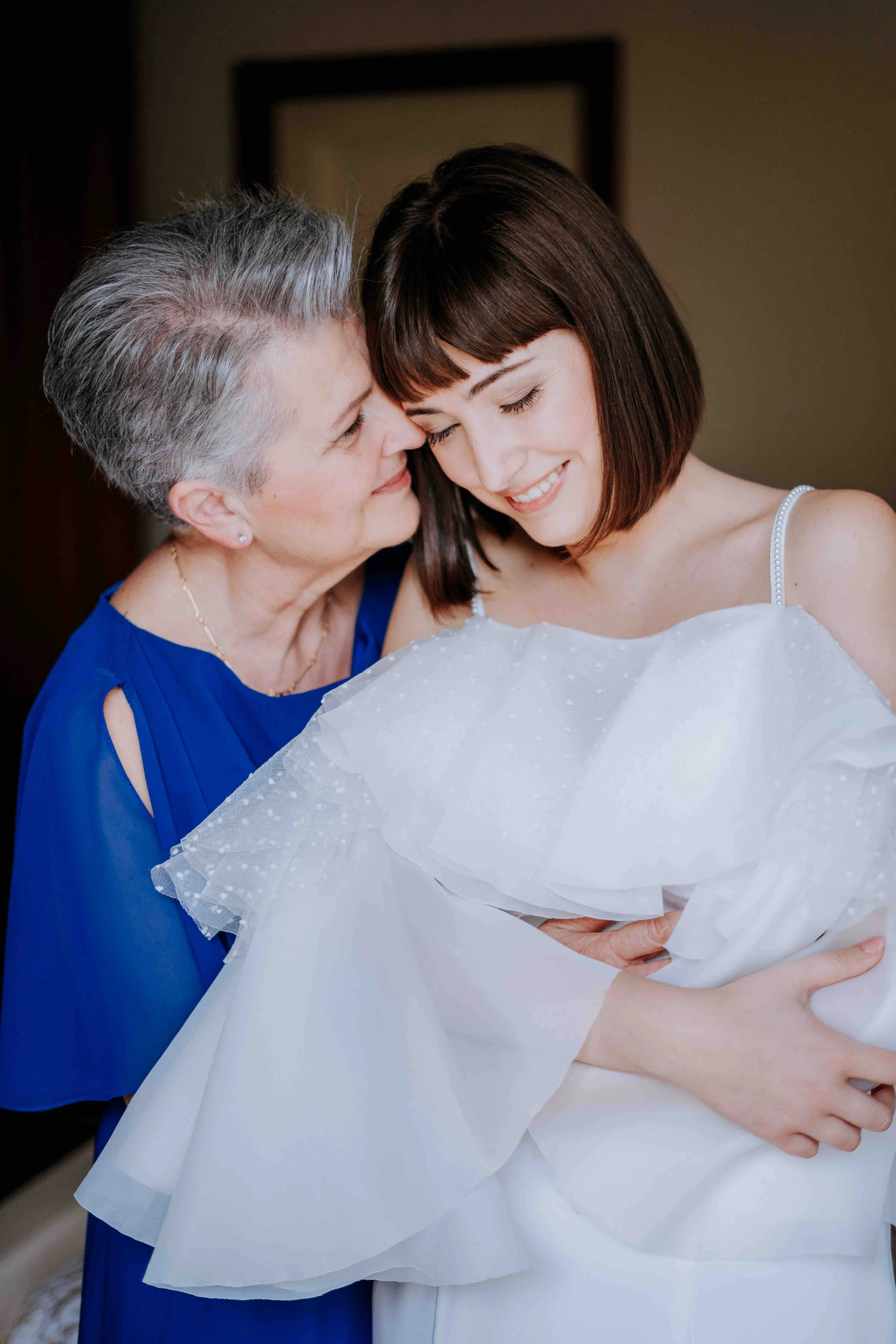 A mother and daughter sharing an intimate moment, with the mother affectionately touching her daughter's forehead and the daughter smiling with her eyes closed, dressed in a white dress with ruffled details.