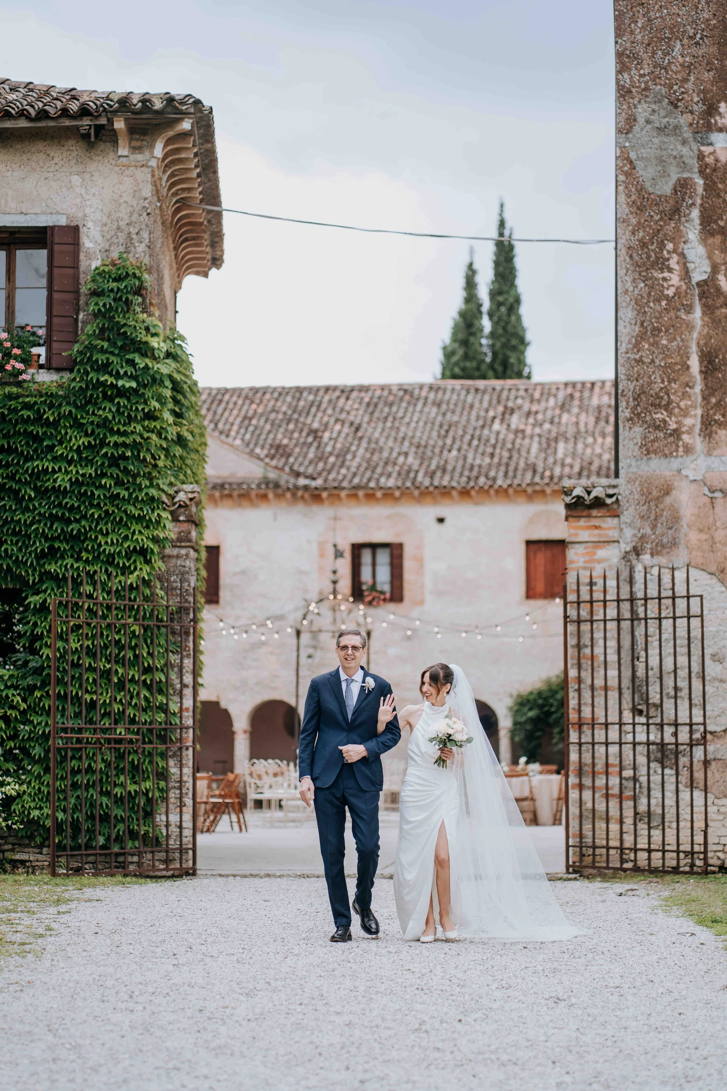 Bride in a white wedding dress holding a bouquet, walking with a man in a blue suit, at a rustic outdoor wedding venue with string lights and historic buildings.