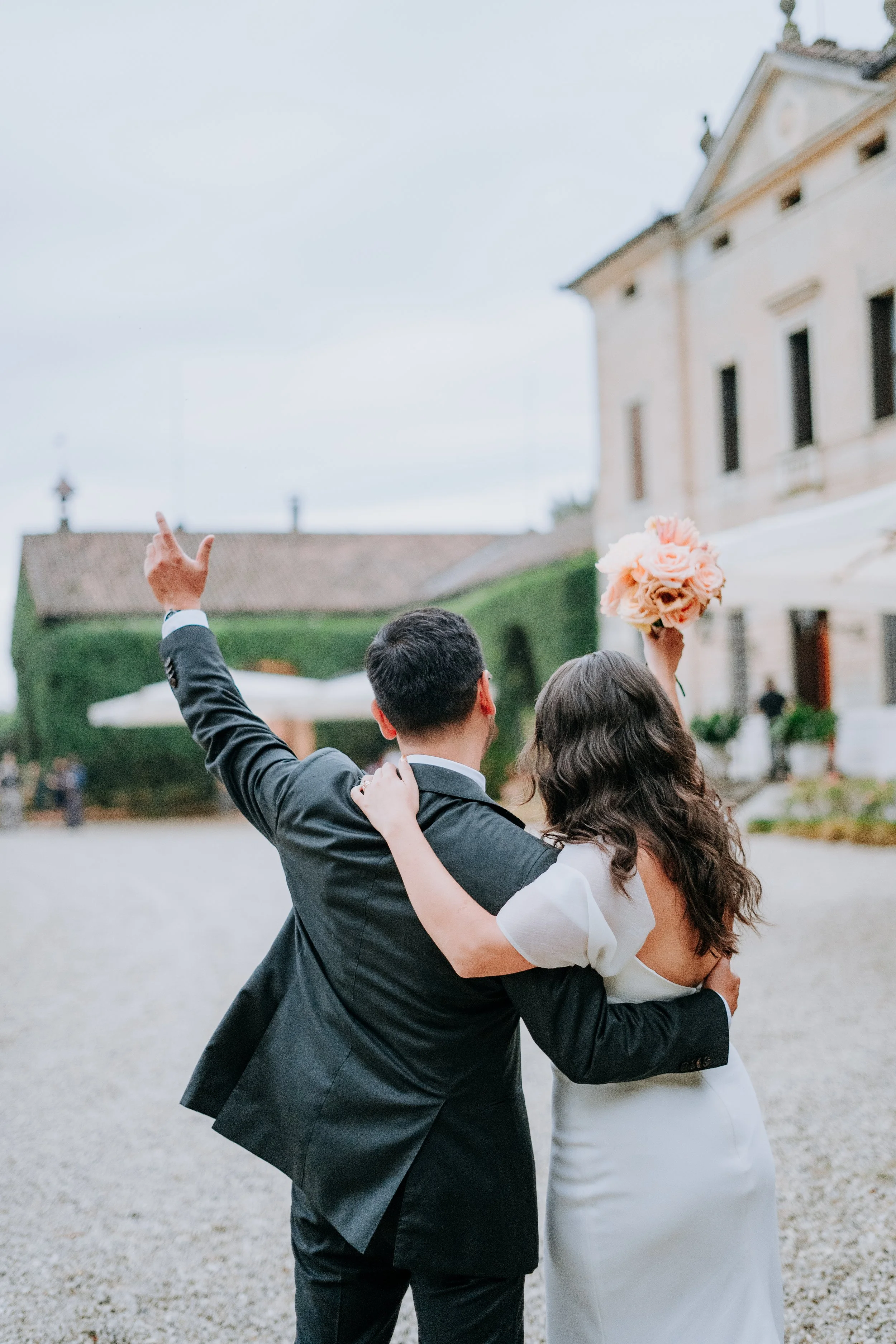 A bride and groom embrace outdoors, with the bride holding a bouquet of pink roses, the groom in a black tuxedo, and the bride in a white wedding dress.