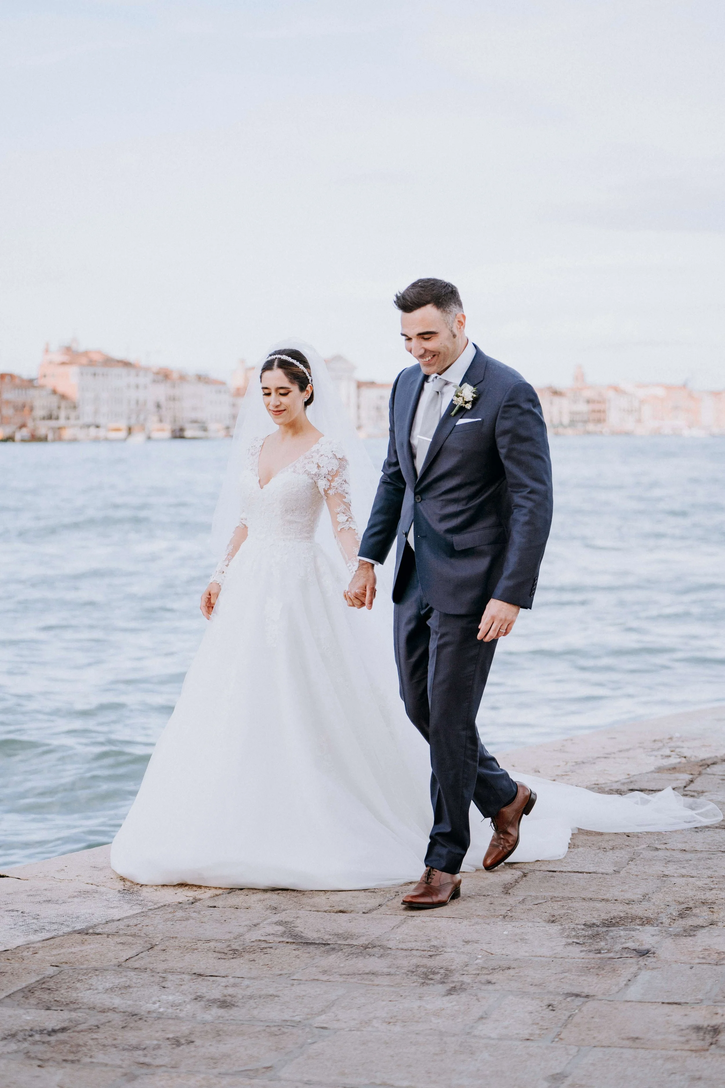 A bride and groom walking hand in hand along a waterfront, dressed in wedding attire, with a cityscape in the background.