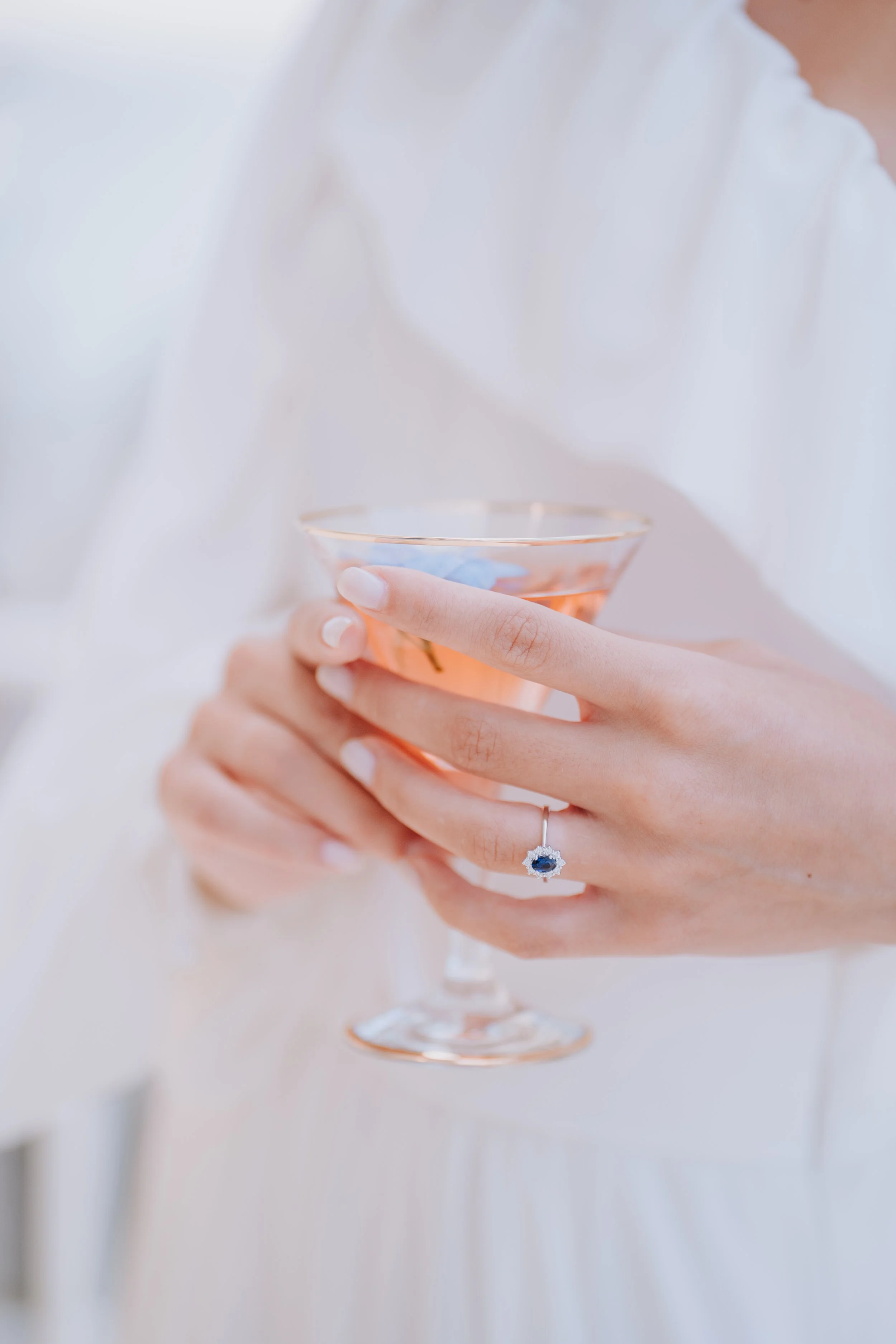 A woman holding a cocktail glass with pink drink, wearing a silver ring with a blue gemstone, and dressed in a white garment.