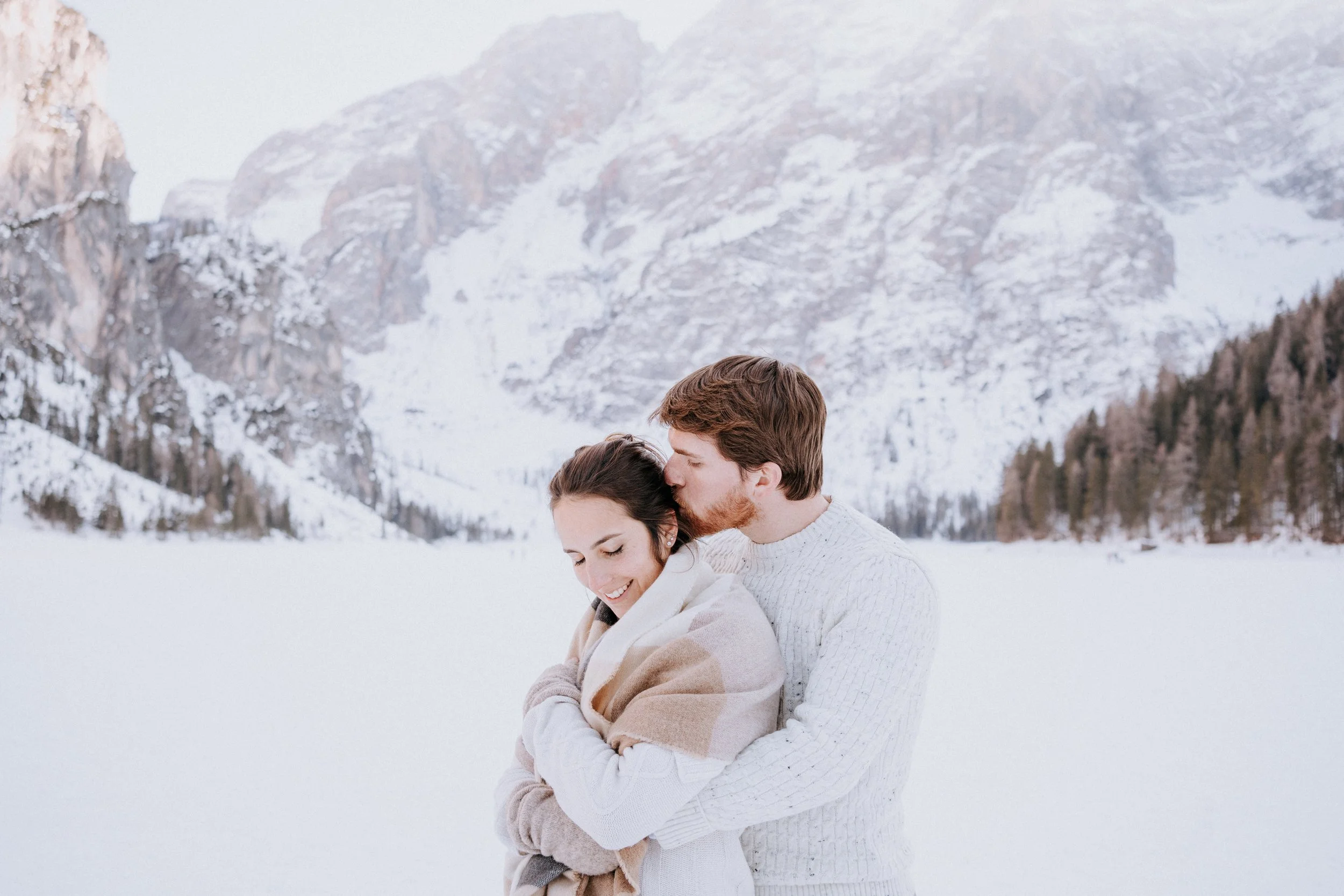 A couple bundled in warm clothing hugging and sharing a kiss outdoors in a snowy mountain landscape.