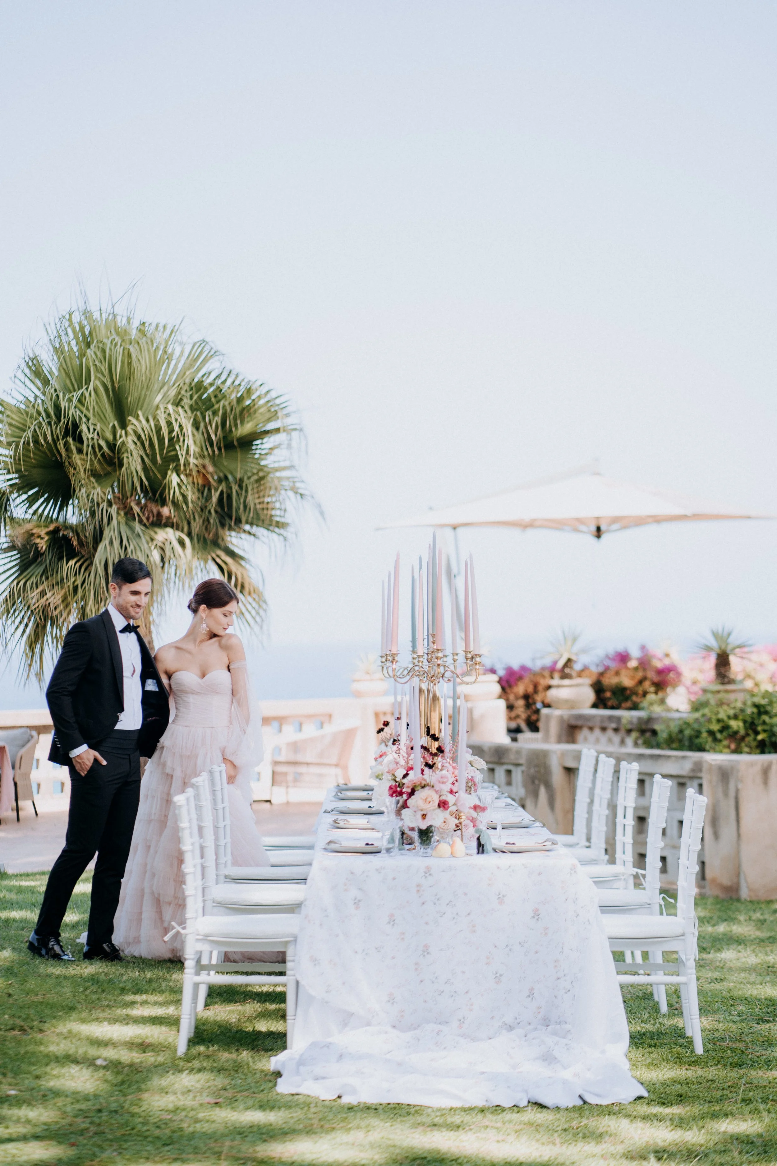 A bride and groom in wedding attire stand beside a decorated outdoor dining table with pink flowers and tall candlesticks, overlooking a scenic view with palm trees and the ocean in the background.